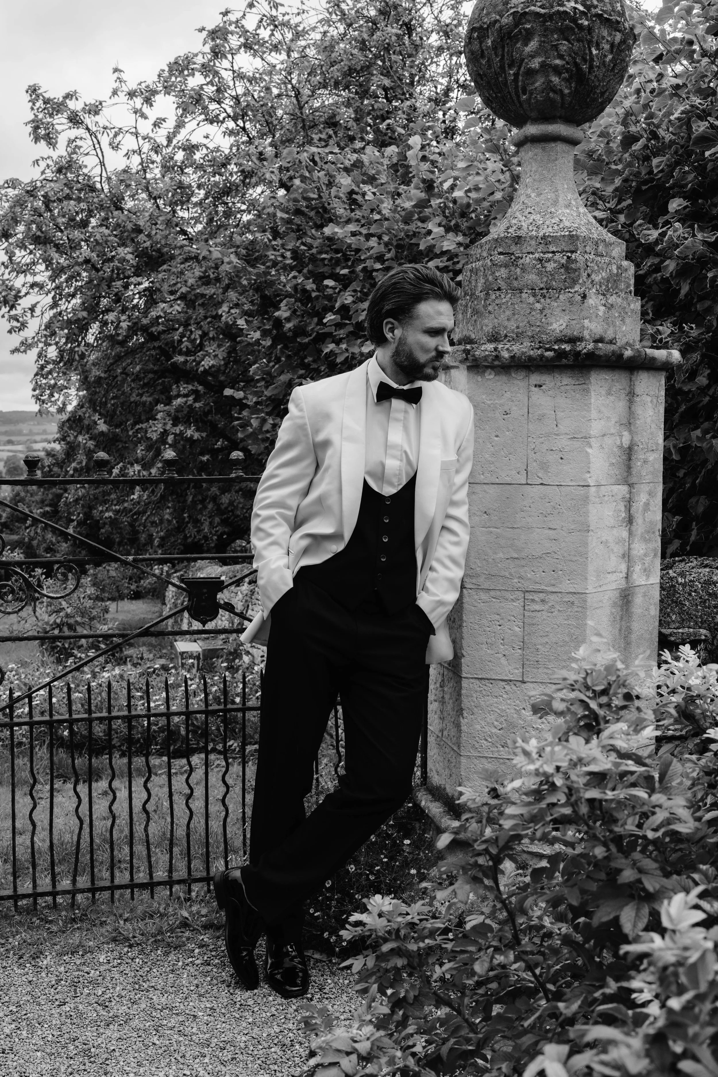 A man in a tuxedo and white blazer leaning against a stone pillar in a garden, looking contemplative. groom prep at euridge manor 