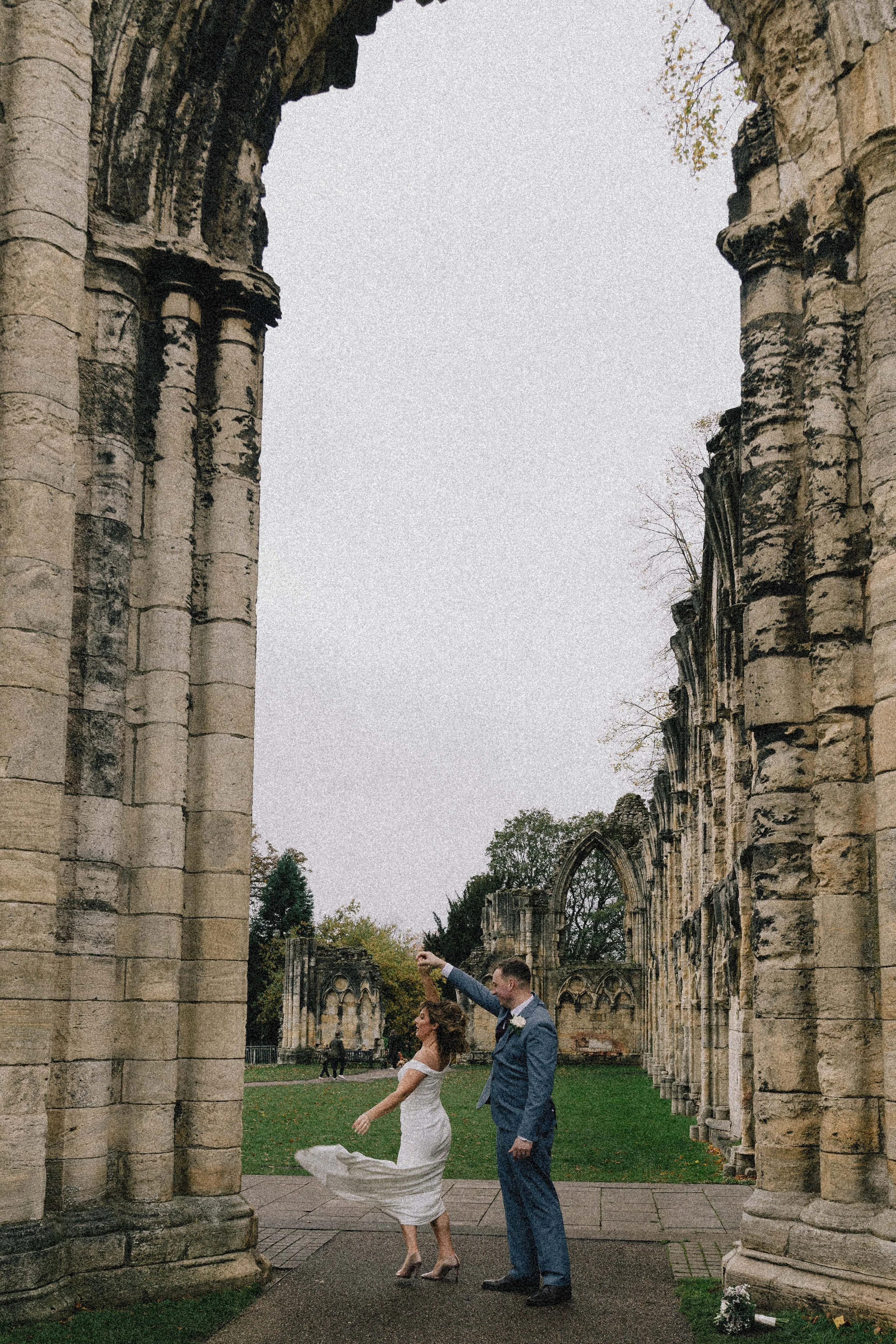 A bride and groom dancing together in the ruins of an old stone building, with an overcast sky overhead.