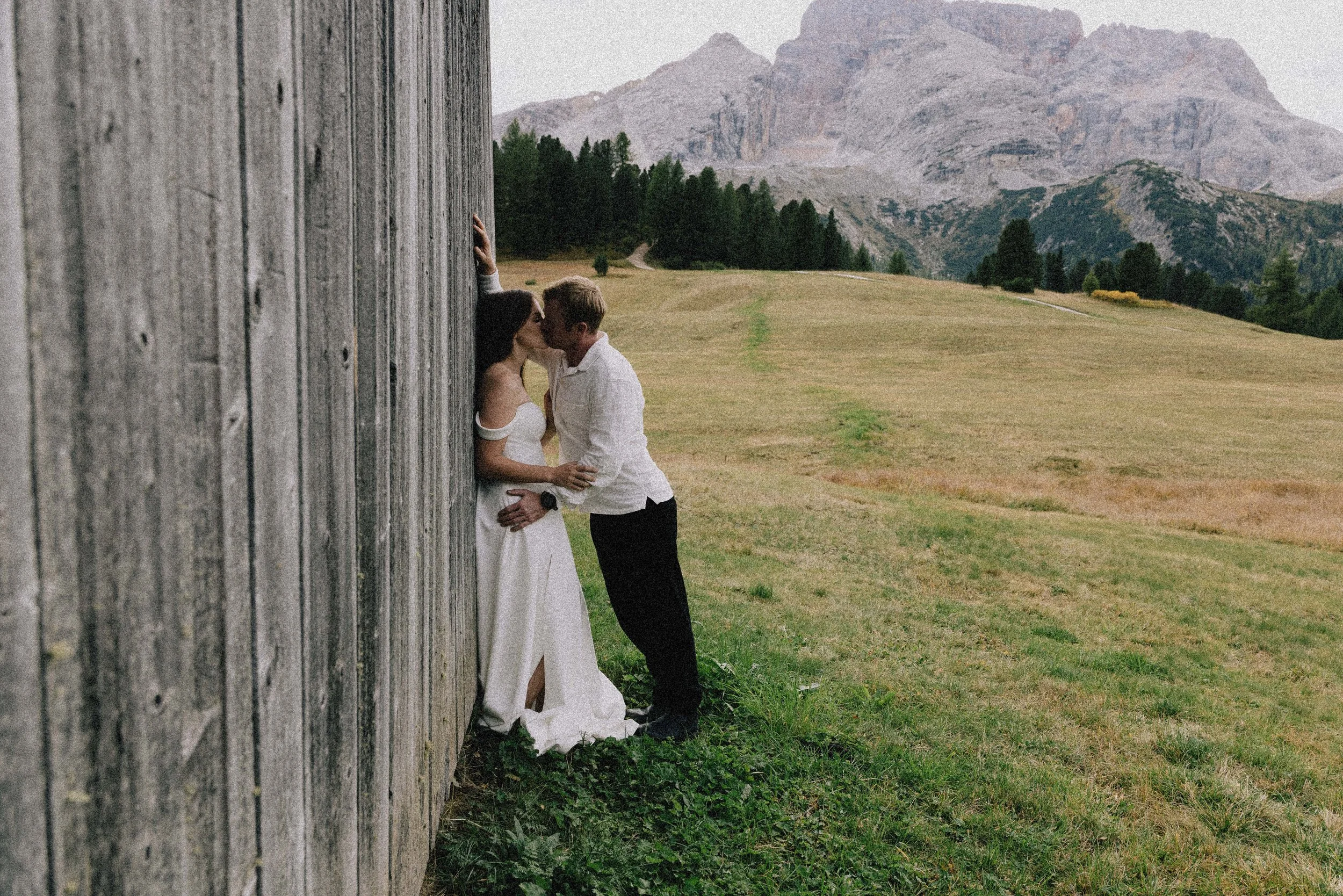 A couple kissing next to a rustic wooden barn in a rural landscape with green fields and mountains in the background.