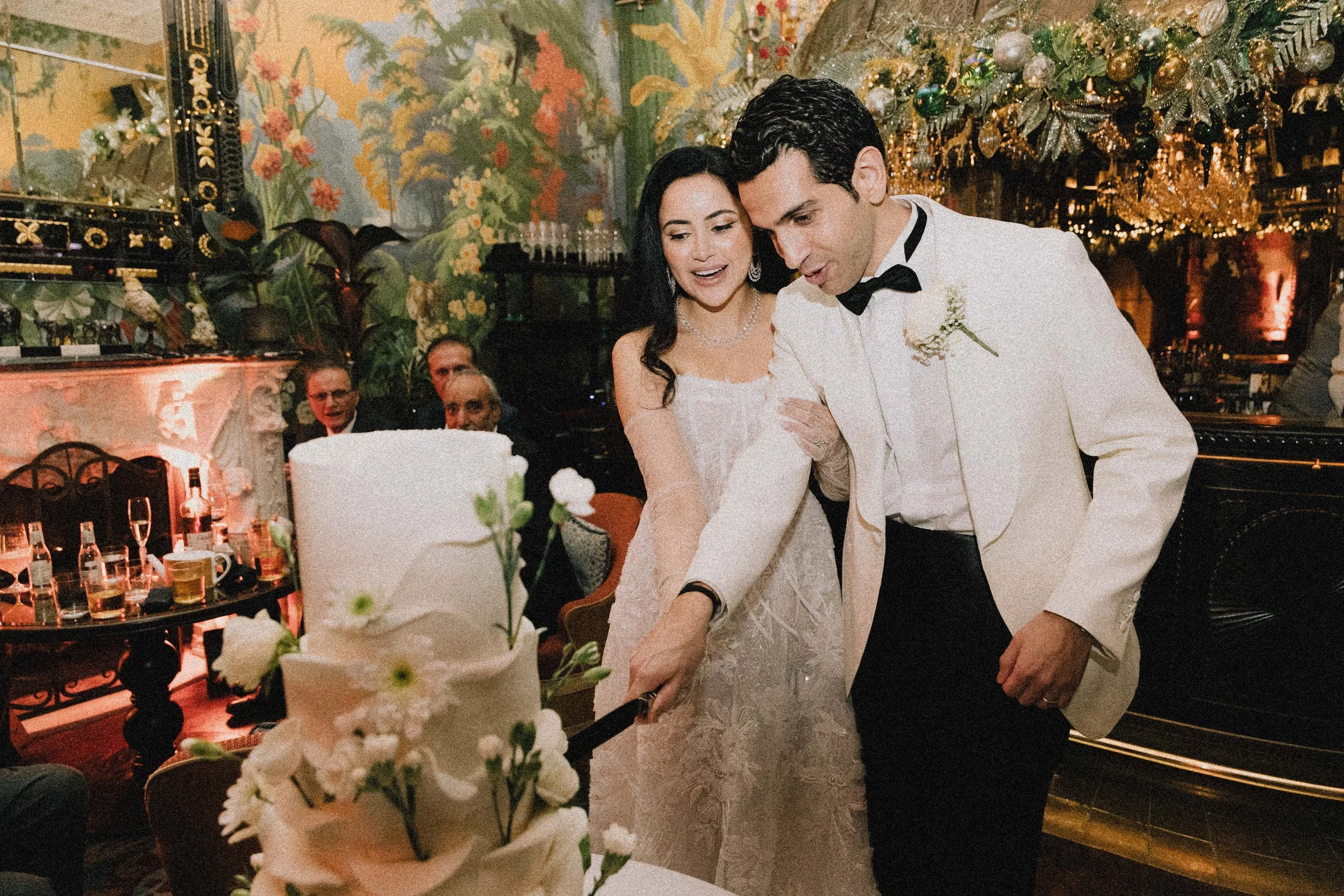 A bride and groom in wedding attire cutting a wedding cake at their celebration, with guests and decorations in the background. Wedding at Anabels in london. London wedding photographer, documentary wedding photography 