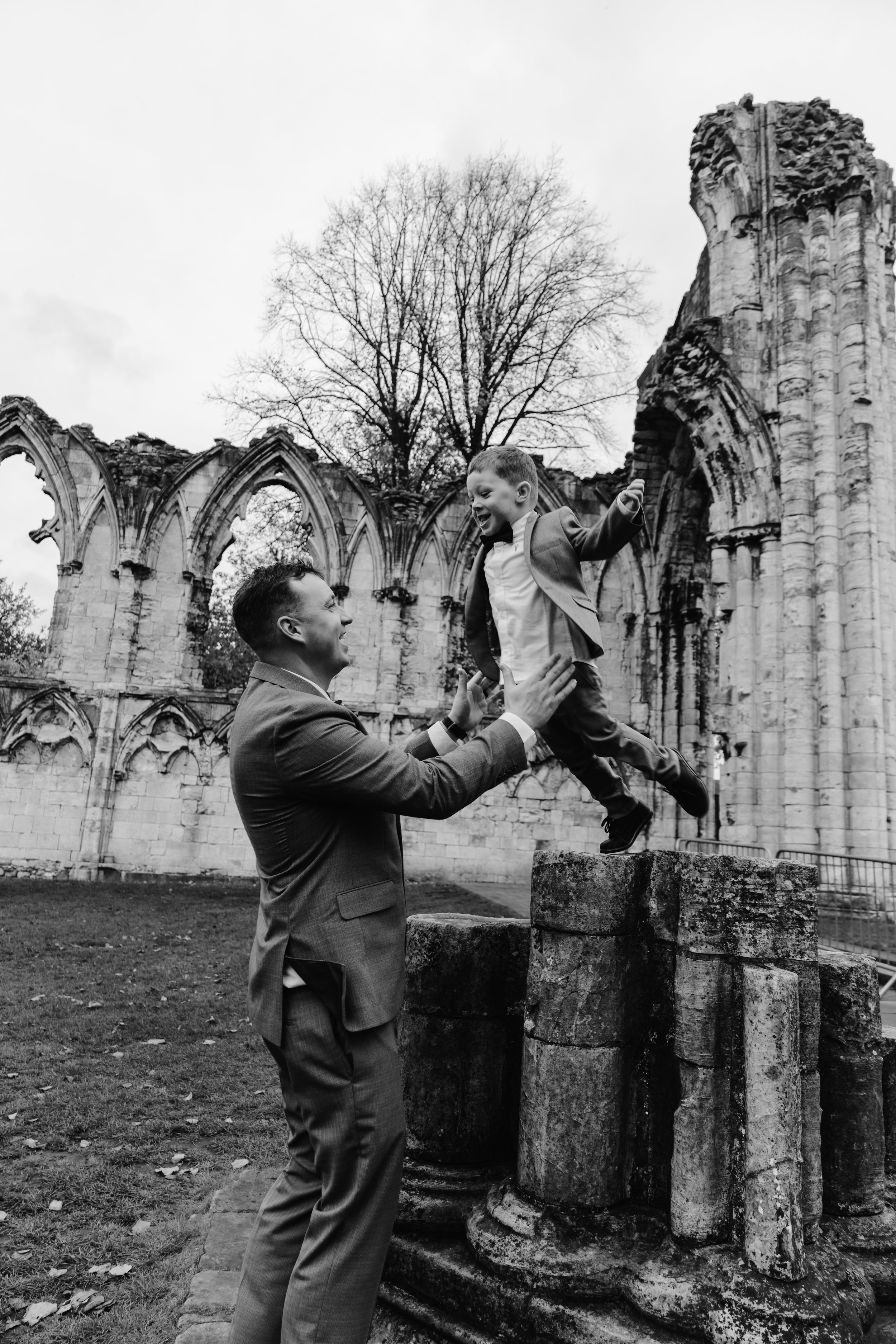 A man and young boy in suits playing on ancient stone ruins outdoors, with some leafless trees in the background. documentary wedding photography, york registry office wedding 