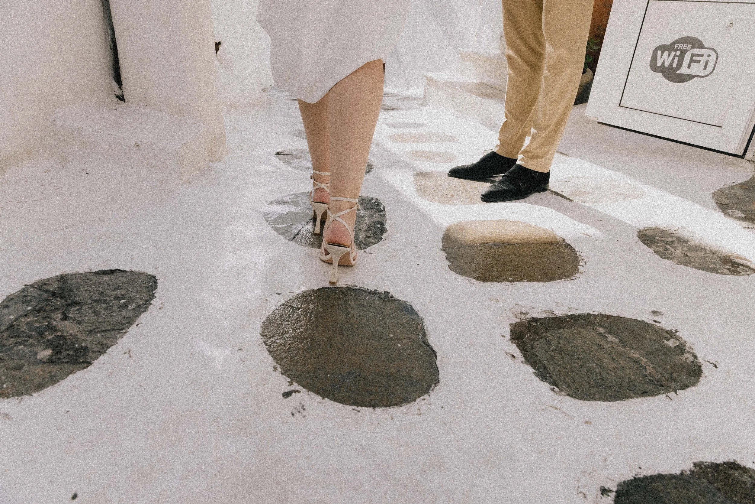A woman in beige high heels and a beige dress walking on a path with large stones, while a man in black shoes and beige pants is standing nearby. There is a sign with Wi-Fi information in the background.