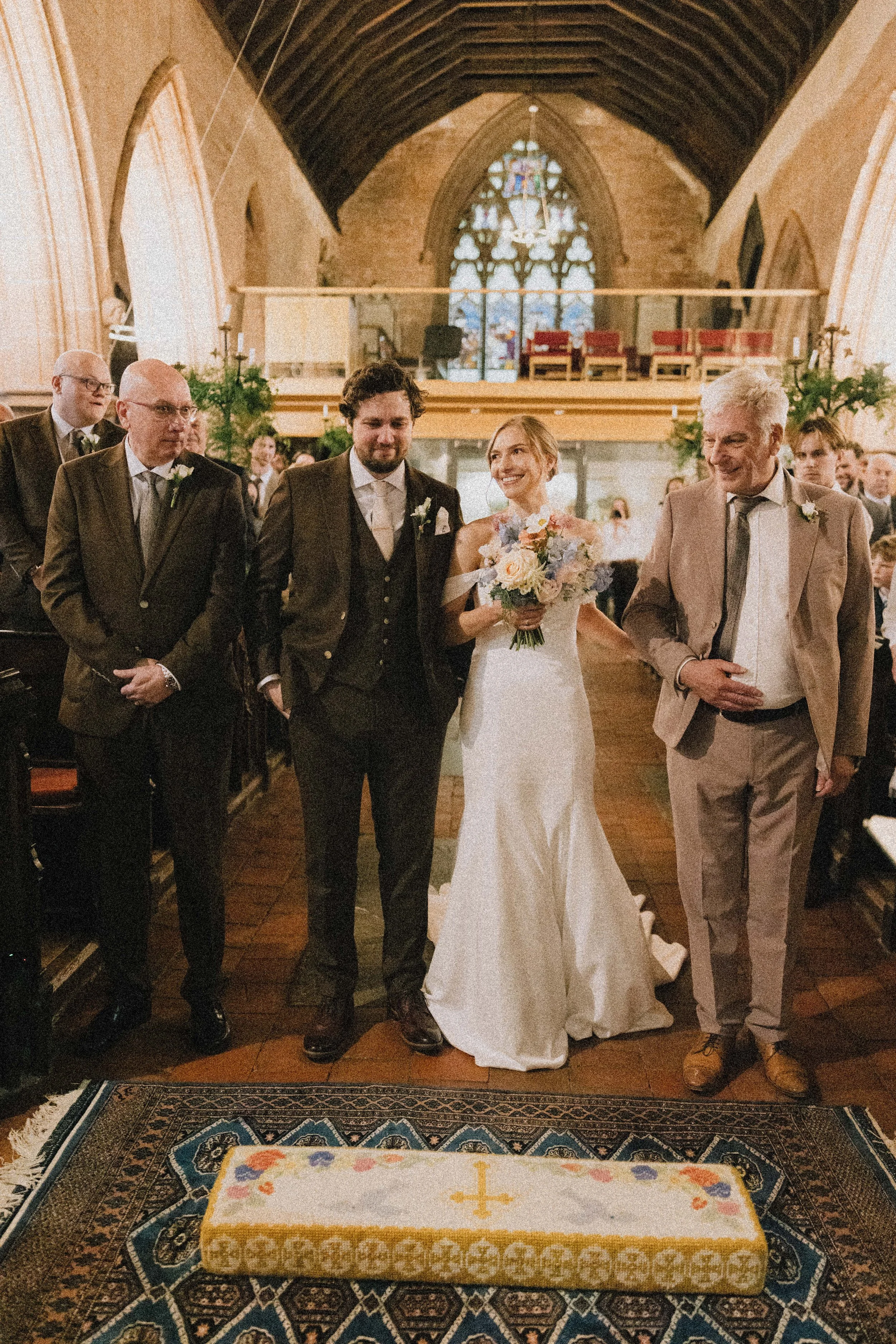 A bride in a white wedding dress holding a bouquet, walking with three men in suits inside a church, with stained glass window in the background and a decorated altar in front.