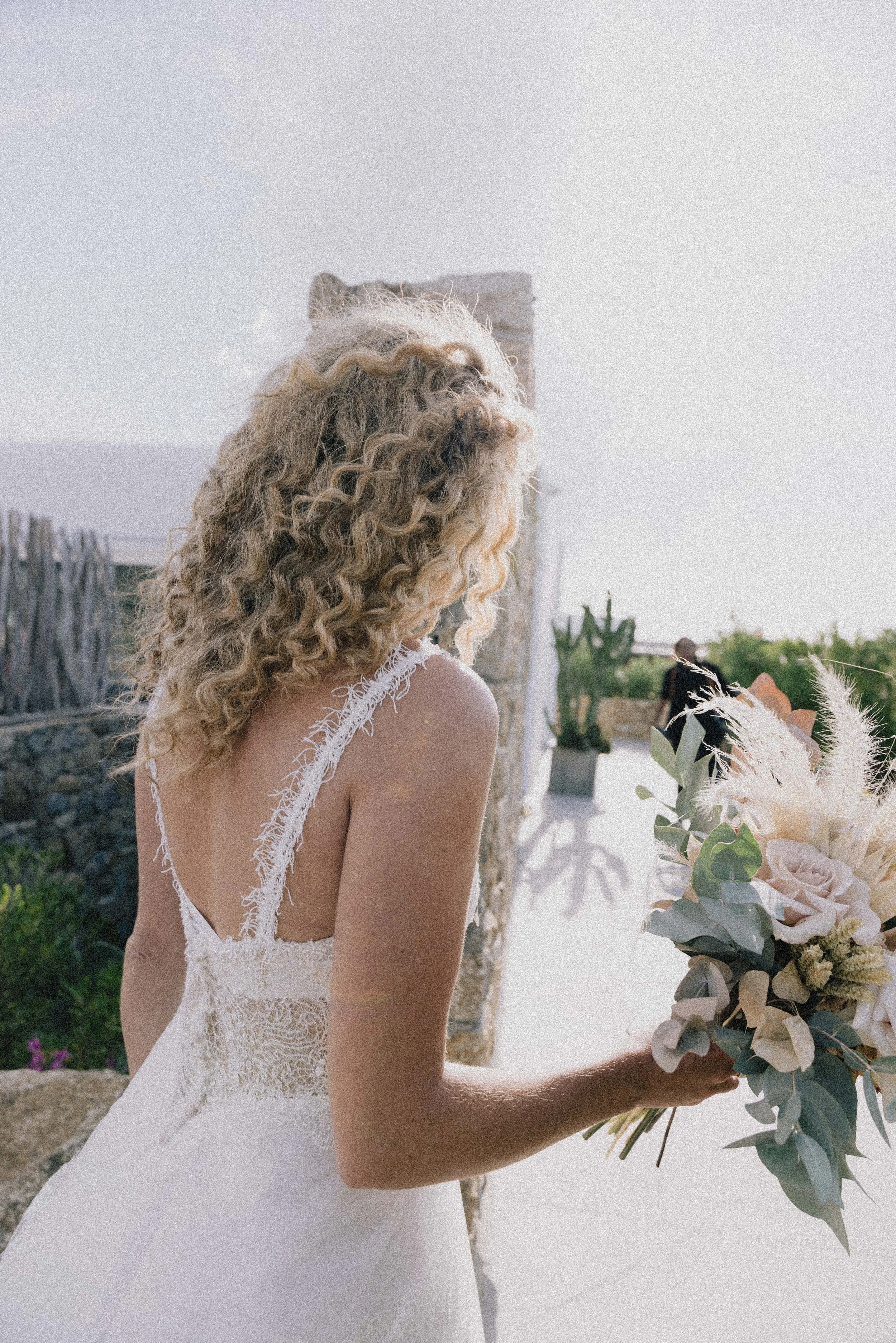 A woman with blonde, curly hair wearing a white wedding dress, holding a bouquet of flowers, standing outdoors with stone and greenery background.
