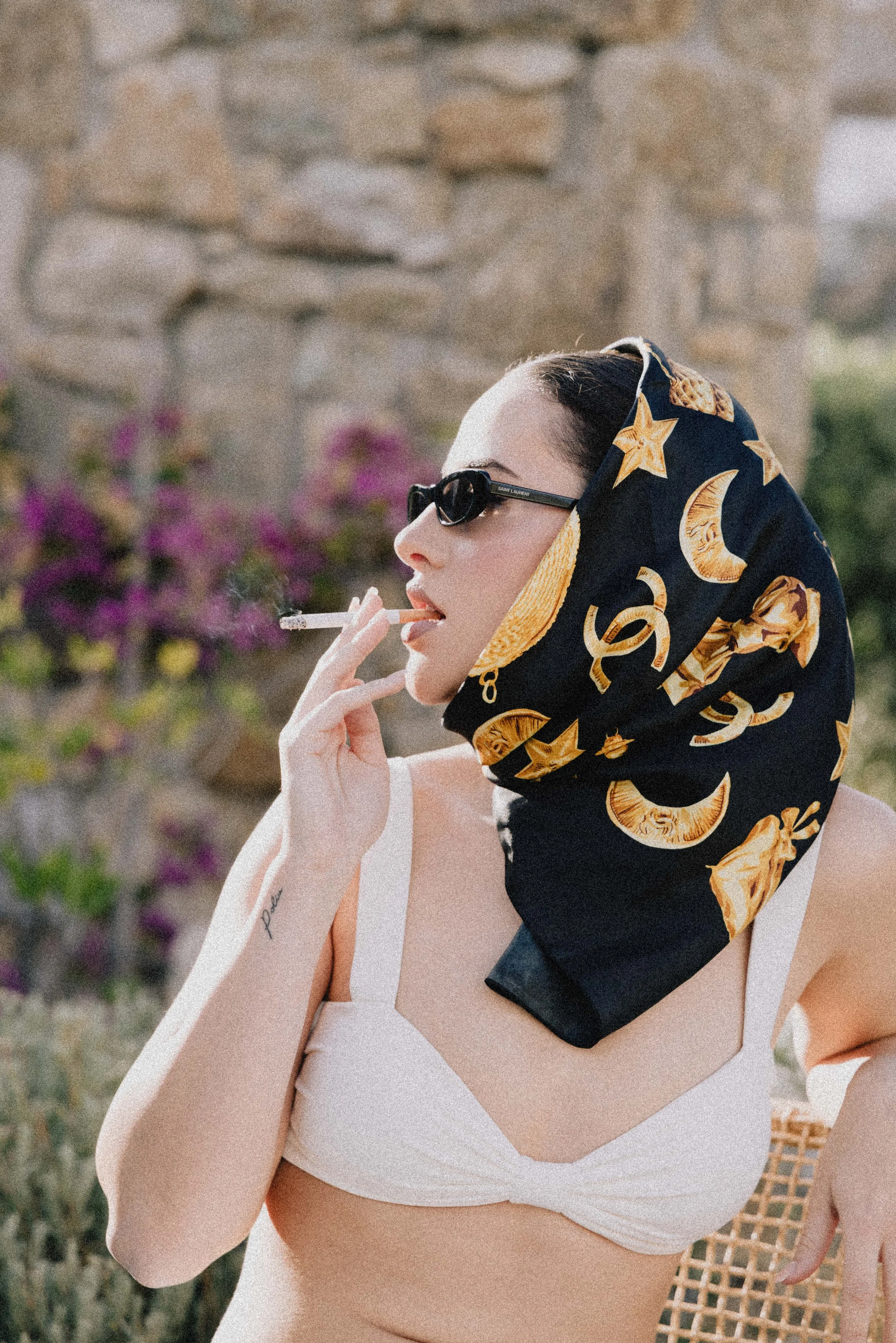 Woman wearing sunglasses and a black headscarf with gold star, moon, and dollar signs pattern, holding a cigarette outdoors near a stone wall.