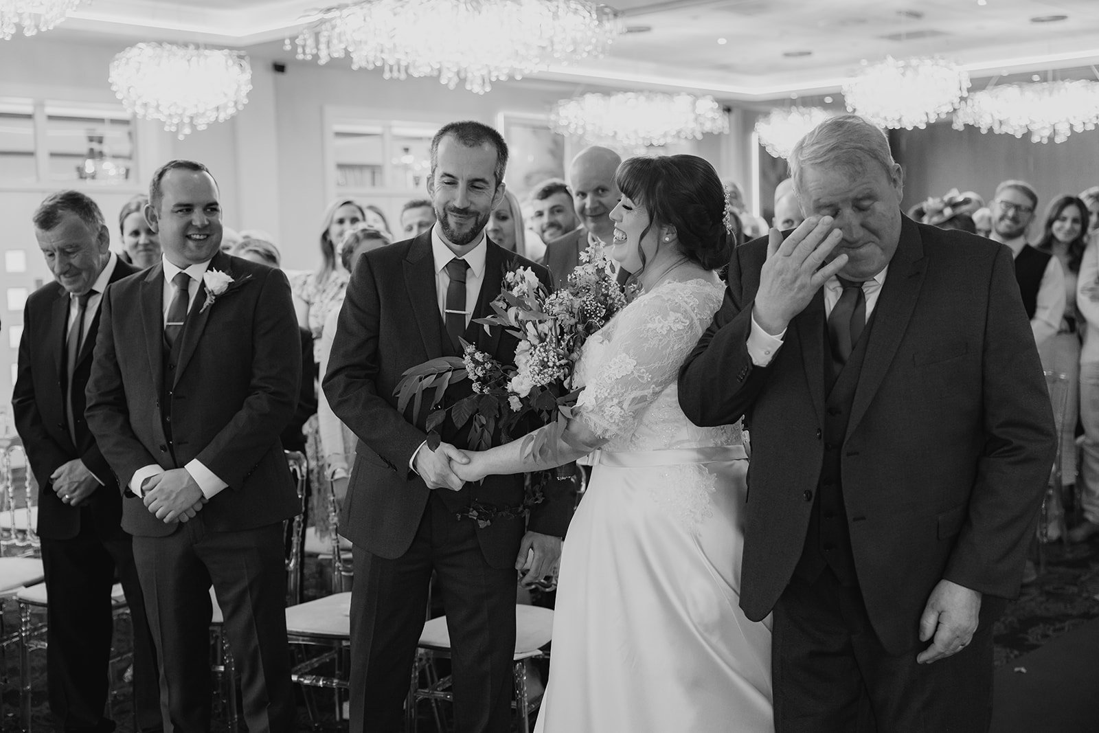 A wedding ceremony with the bride and groom holding hands, surrounded by emotional guests in a reception hall with chandeliers. cheshire wedding venue thorton hall