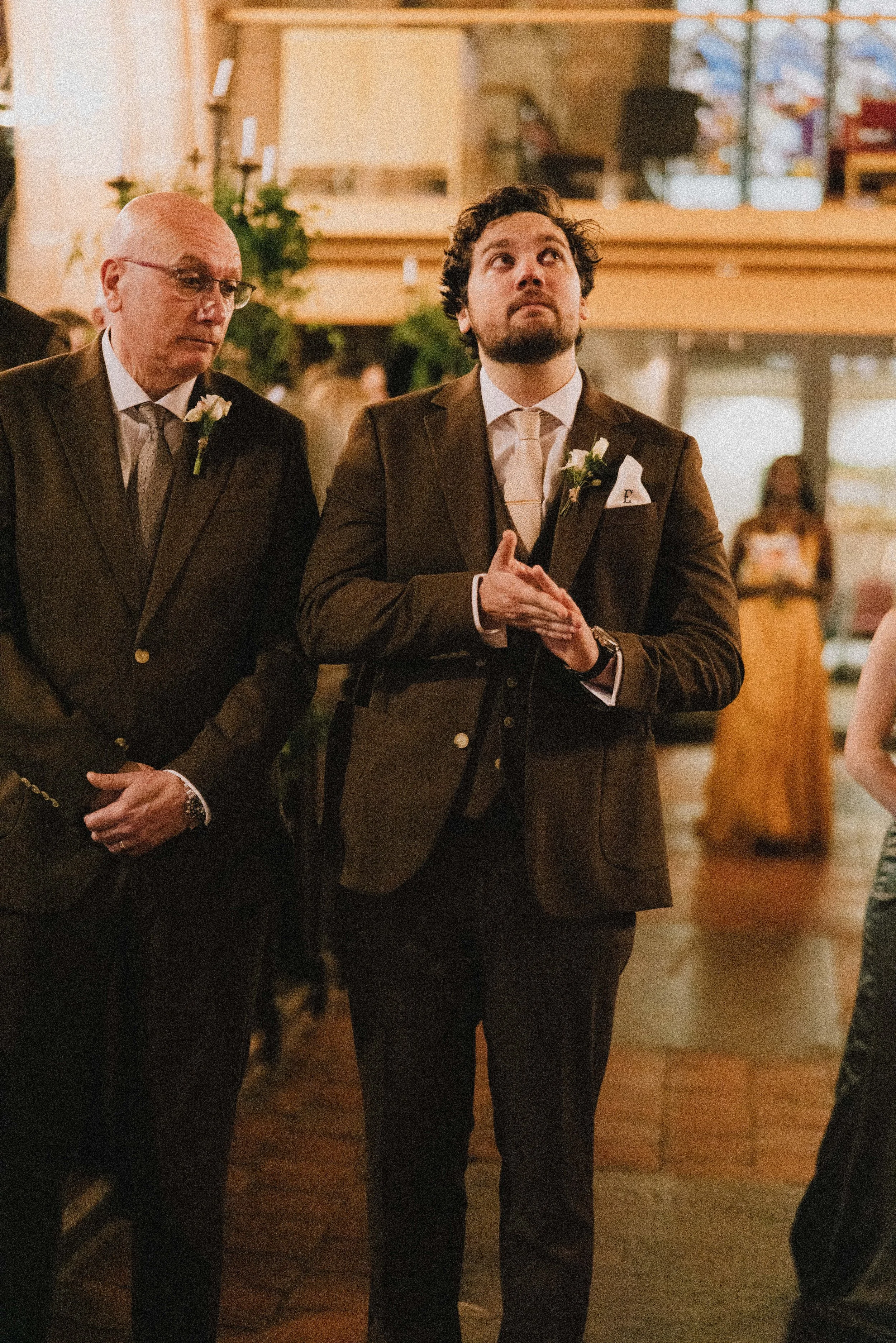 A man wearing a brown suit, white shirt, and light-colored tie, standing with his hands clasped in a prayerful position, in a formal indoor setting, possibly a wedding, with an older man in a similar suit standing to his left.