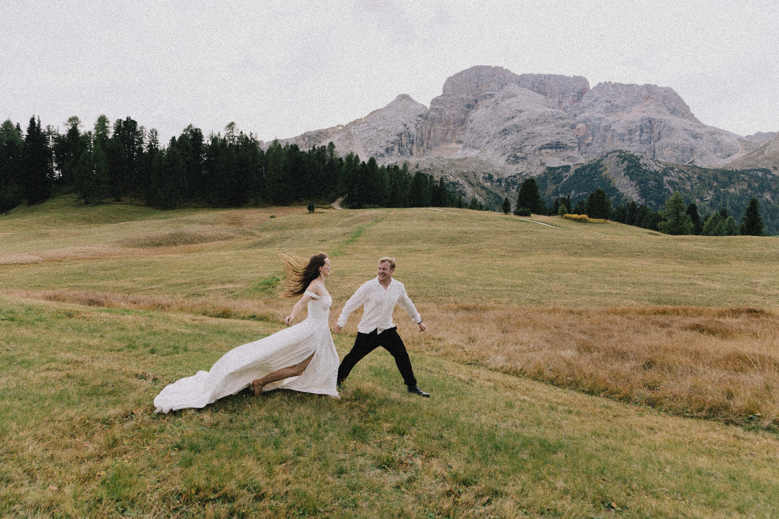 A woman in a white wedding dress running hand-in-hand with a man in a white shirt and black pants across a grassy field with mountains in the background.