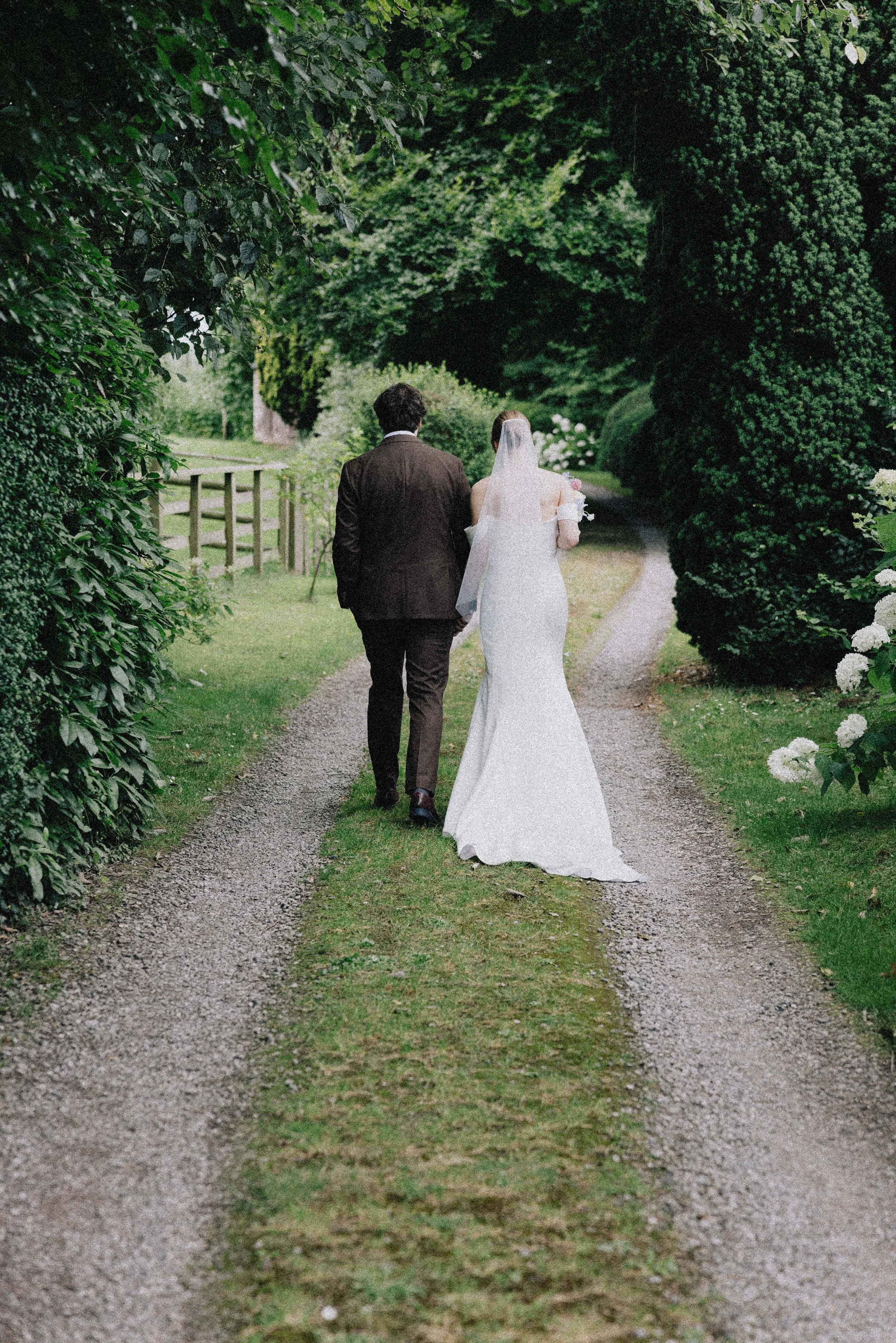 A bride and groom walking hand in hand down a gravel path in a lush, green garden or park.