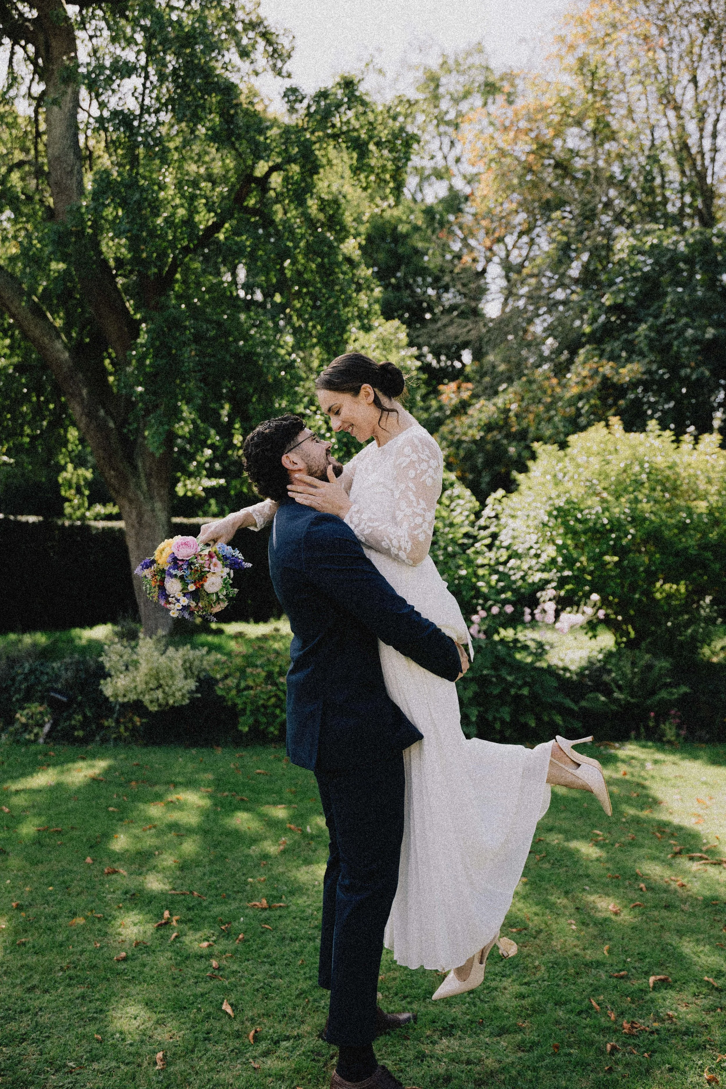 A happy couple, dressed in wedding attire, embrace in a park-like setting with trees and greenery, one lifting the other in celebration.