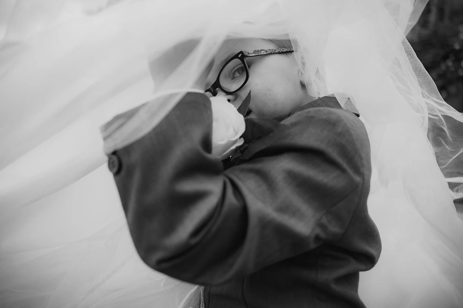 A young girl with glasses and face paint, dressed in a jacket, is lying down wrapped in a sheer fabric, looking to the side in a black-and-white photo. Merrydale Manor Wedding Venue weddings 