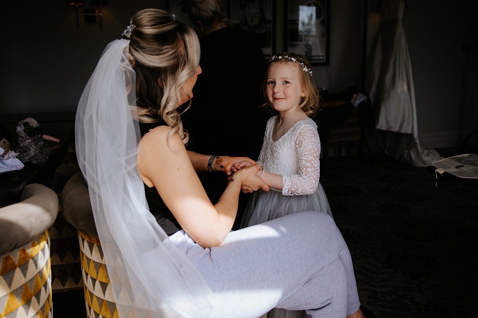 A bride with a wedding veil holding hands with a young girl wearing a lace dress and a floral headband, inside a dimly lit room. wedding at walton hall garden 