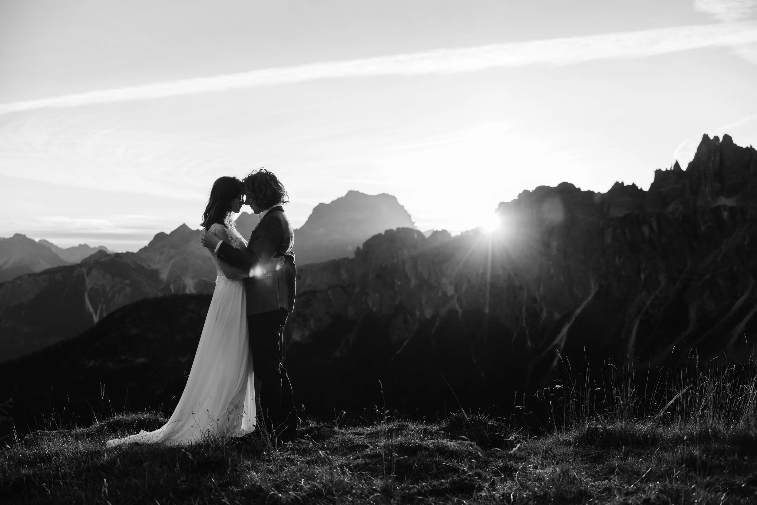 A black-and-white photo of a couple embracing and touching foreheads on a mountain ridge at sunset, with mountain peaks and a bright sun in the background. documentary wedding photographer shooting a wedding in italy 