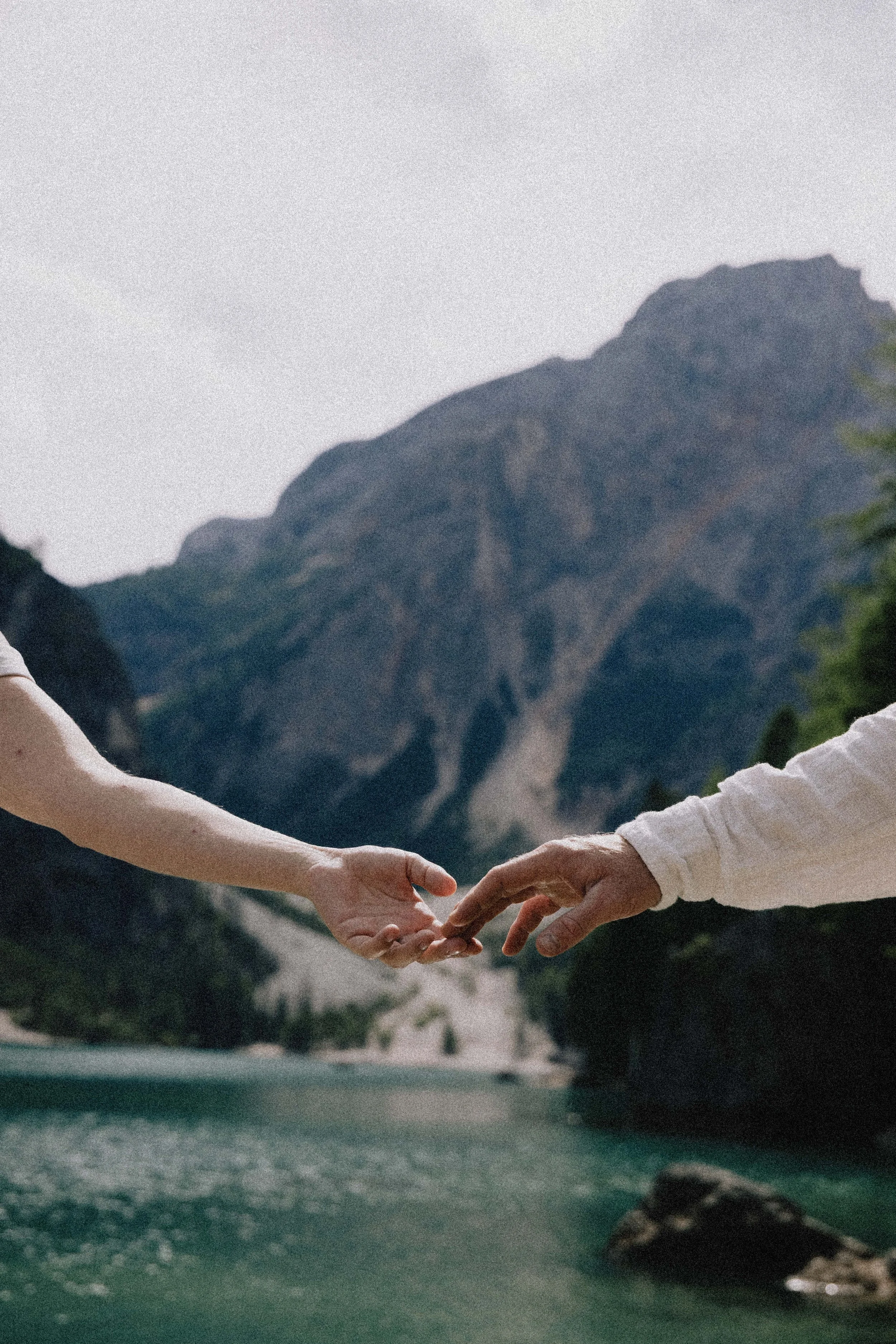 Two people reaching out to hold hands near a mountain lake