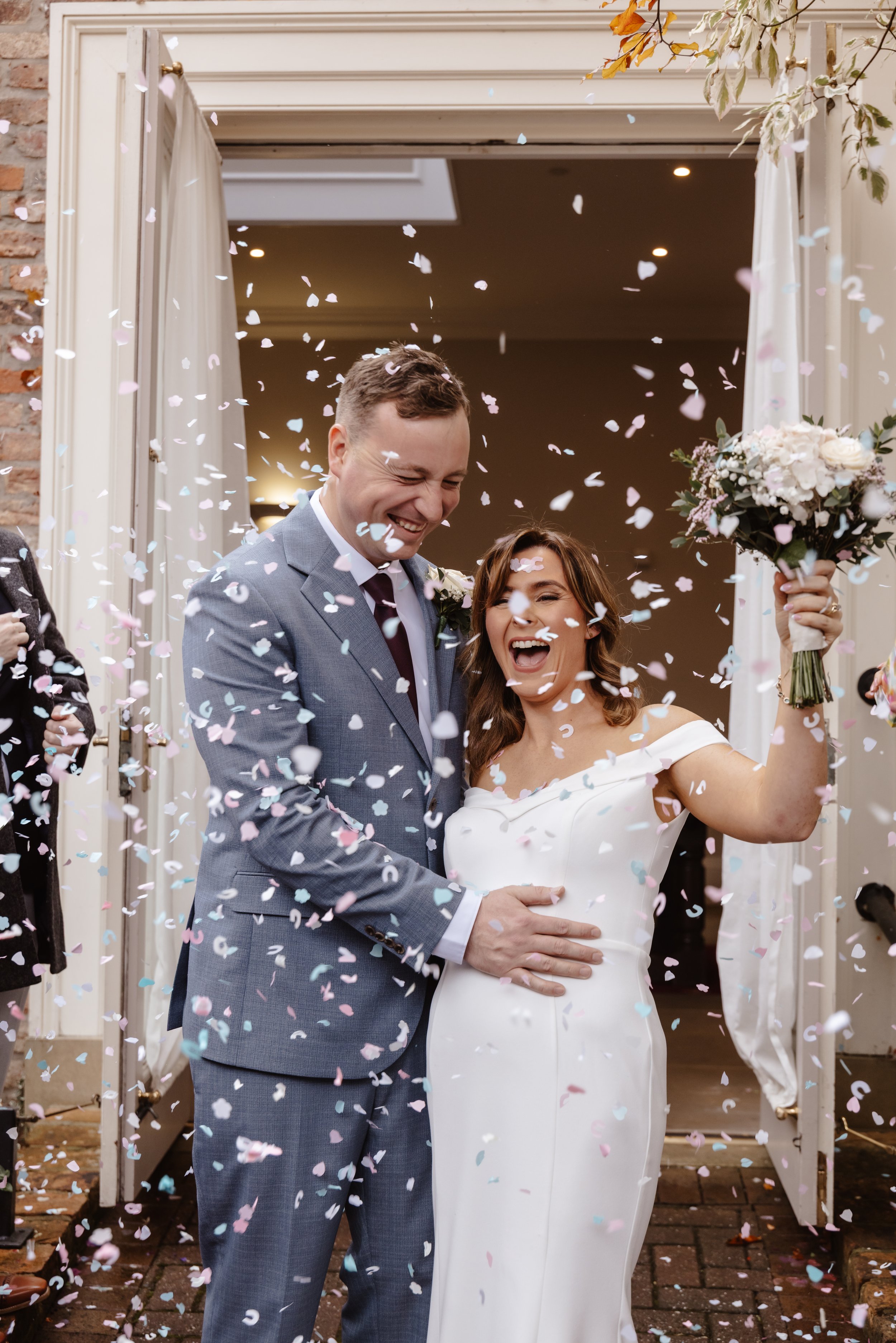 A newlywed couple celebrating as they exit a building, surrounded by falling confetti. The groom is wearing a gray suit with a black tie, and the bride is in a white wedding dress holding a bouquet of flowers. wedding at york registry office