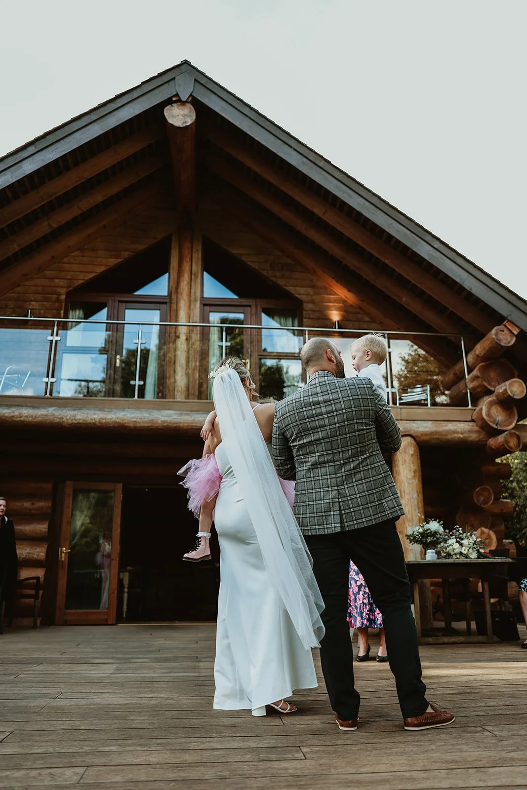 Bride with long veil and white dress standing on wooden deck, holding a young girl in pink dress, in front of a log cabin with large windows and balcony, during a wedding celebration outdoors. hidden river cabin elopements 