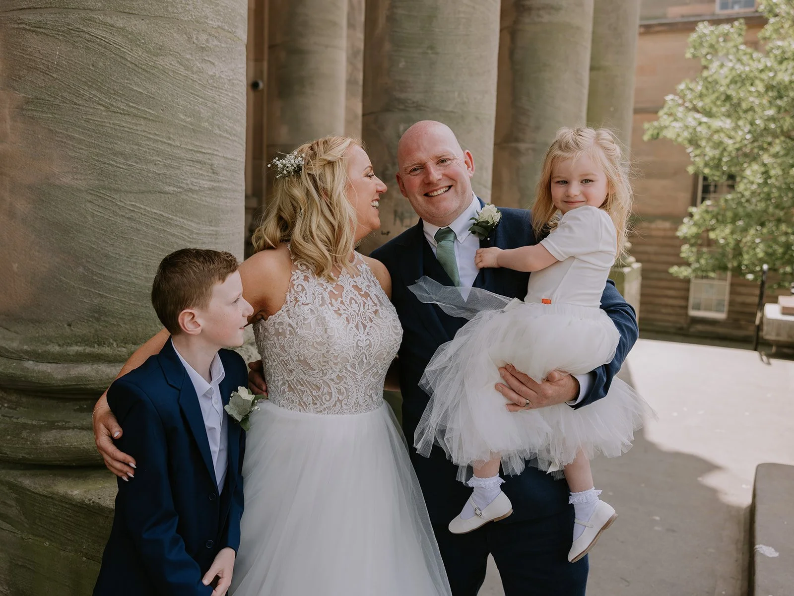 A group of four people, a woman in a wedding dress, a man in a suit, a young girl holding onto the man, and a young boy in a suit, standing outdoors in front of large stone columns, smiling and sharing a joyful moment. cheshire wedding venue 