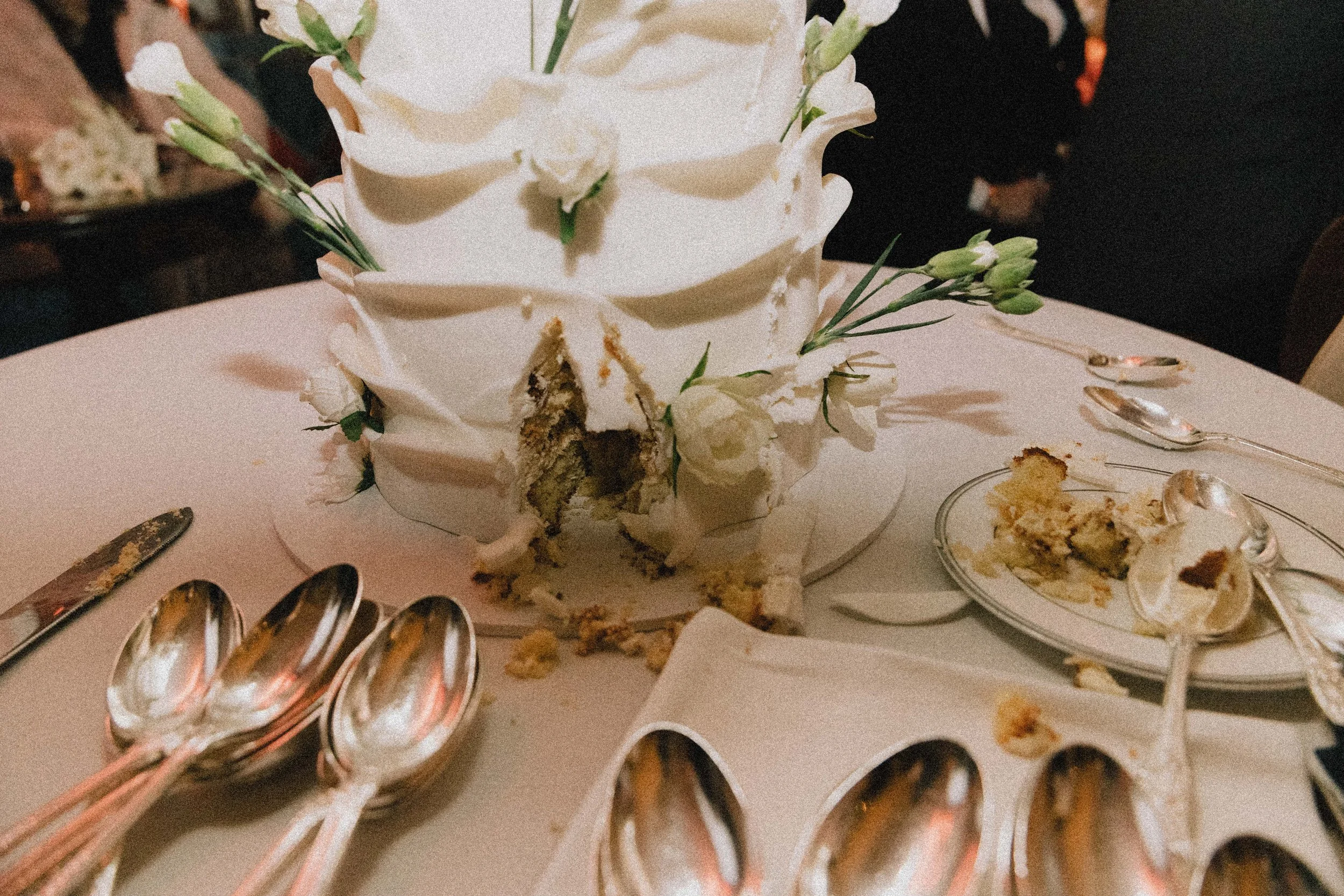 A white wedding cake with flowers, partially cut into, on a table with silver spoons and plates, some cake remnants on the plates and table.