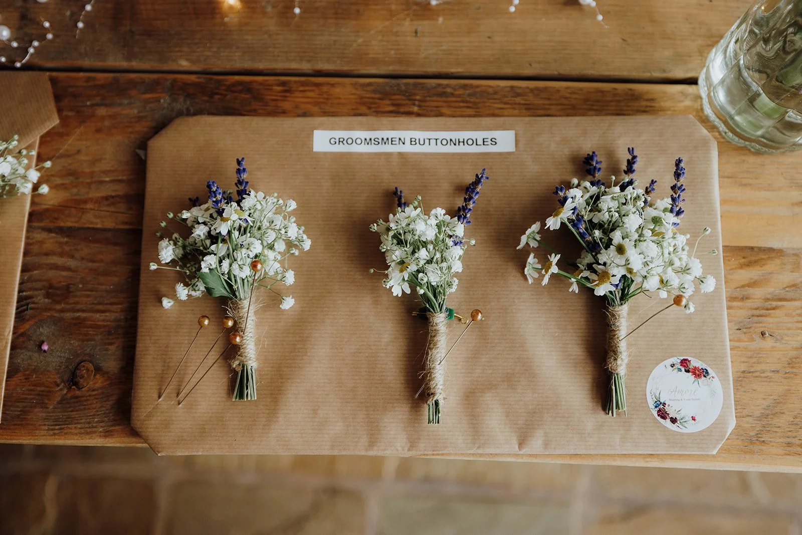 Three boutonniere arrangements of small white flowers with purple sprigs, wrapped with twine, labeled 'GROOMSMEN BUTTONHOLES' on brown paper, on a wooden table.