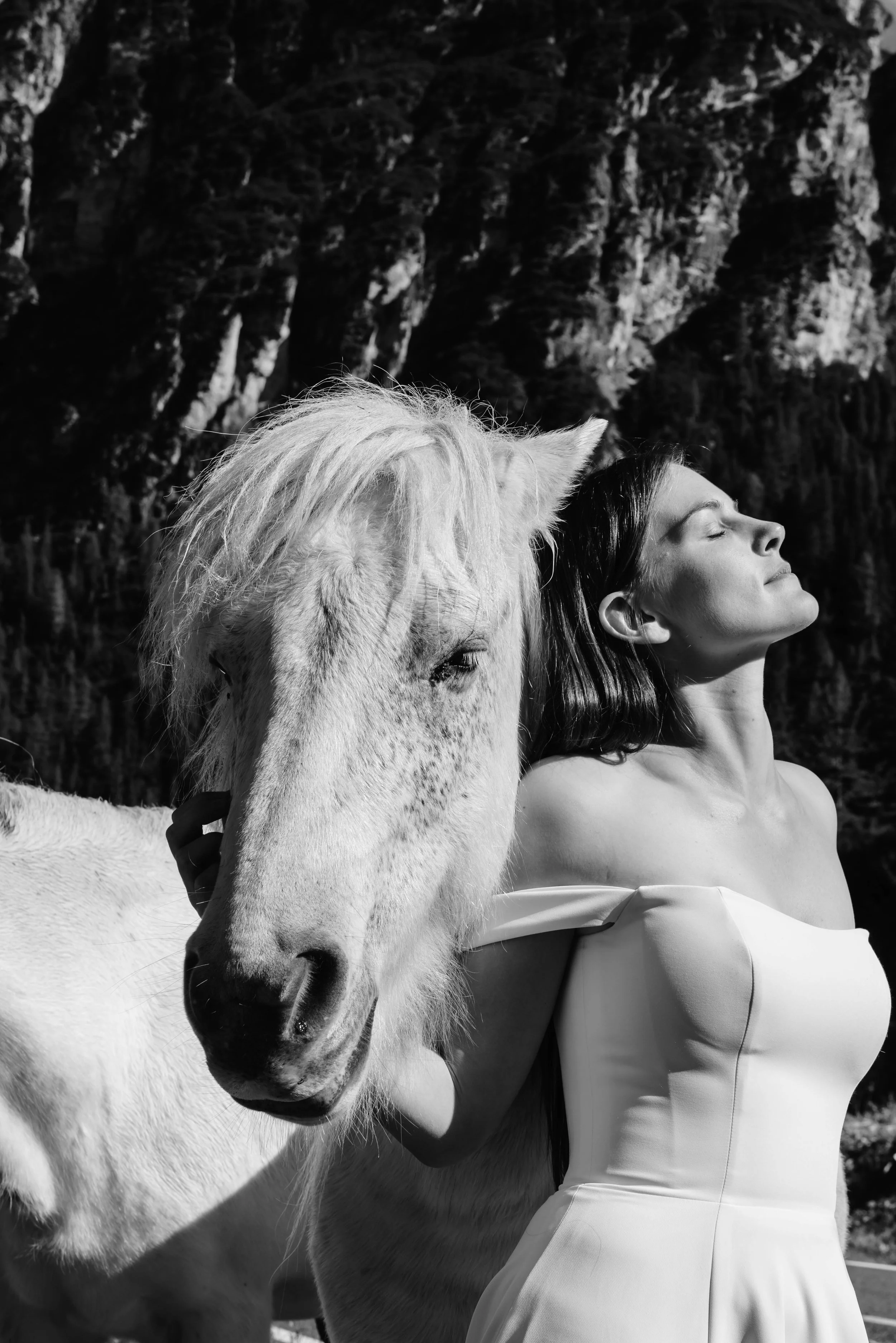 A woman with dark hair and light skin in a strapless dress standing beside a white horse with a light mane, outdoors with rocky cliffs in the background, in black and white. destination wedding photographer in dolomites 