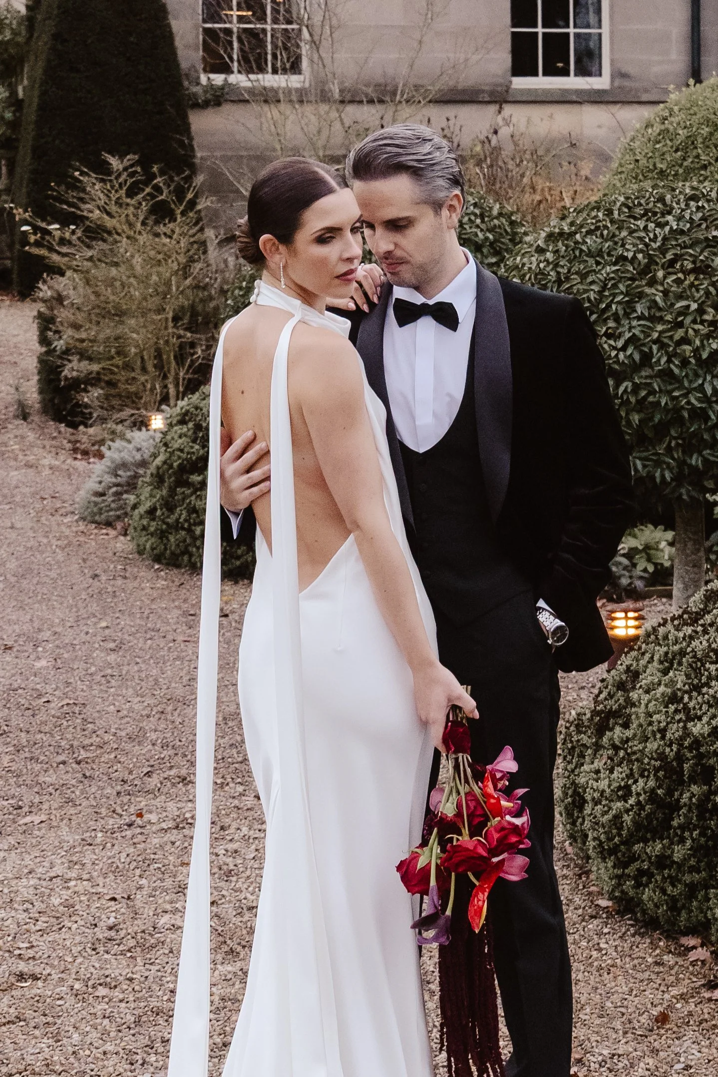 A bride and groom in elegant wedding attire standing close together outdoors, with the bride holding a bouquet of red and purple flowers. an editorial shot of wedding couple at middleton lodge 