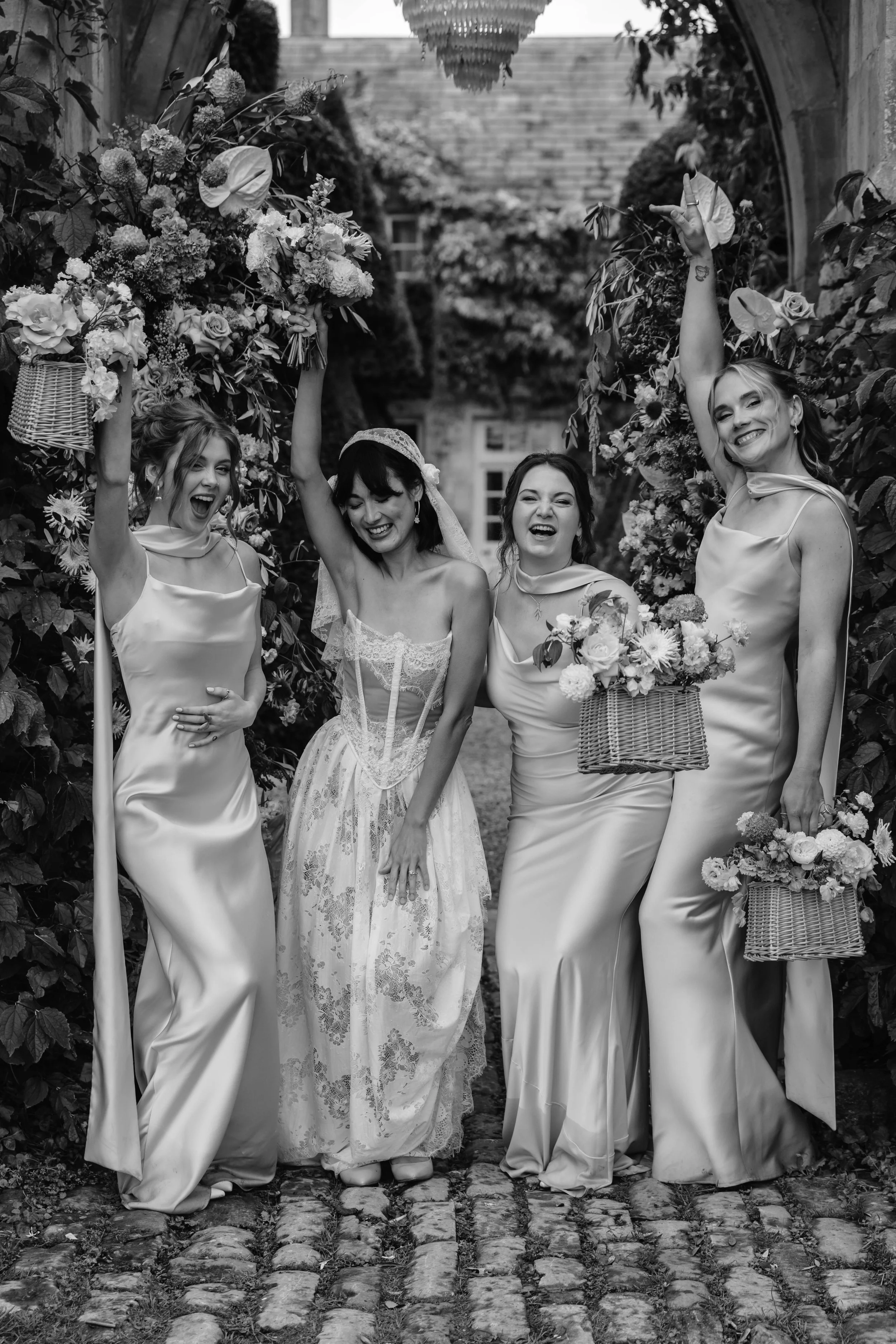 Four women celebrating outdoors, with two holding large bouquets of flowers and baskets, dressed in elegant dresses. documentary wedding photography at the iconic wedding venue Euridge Manor