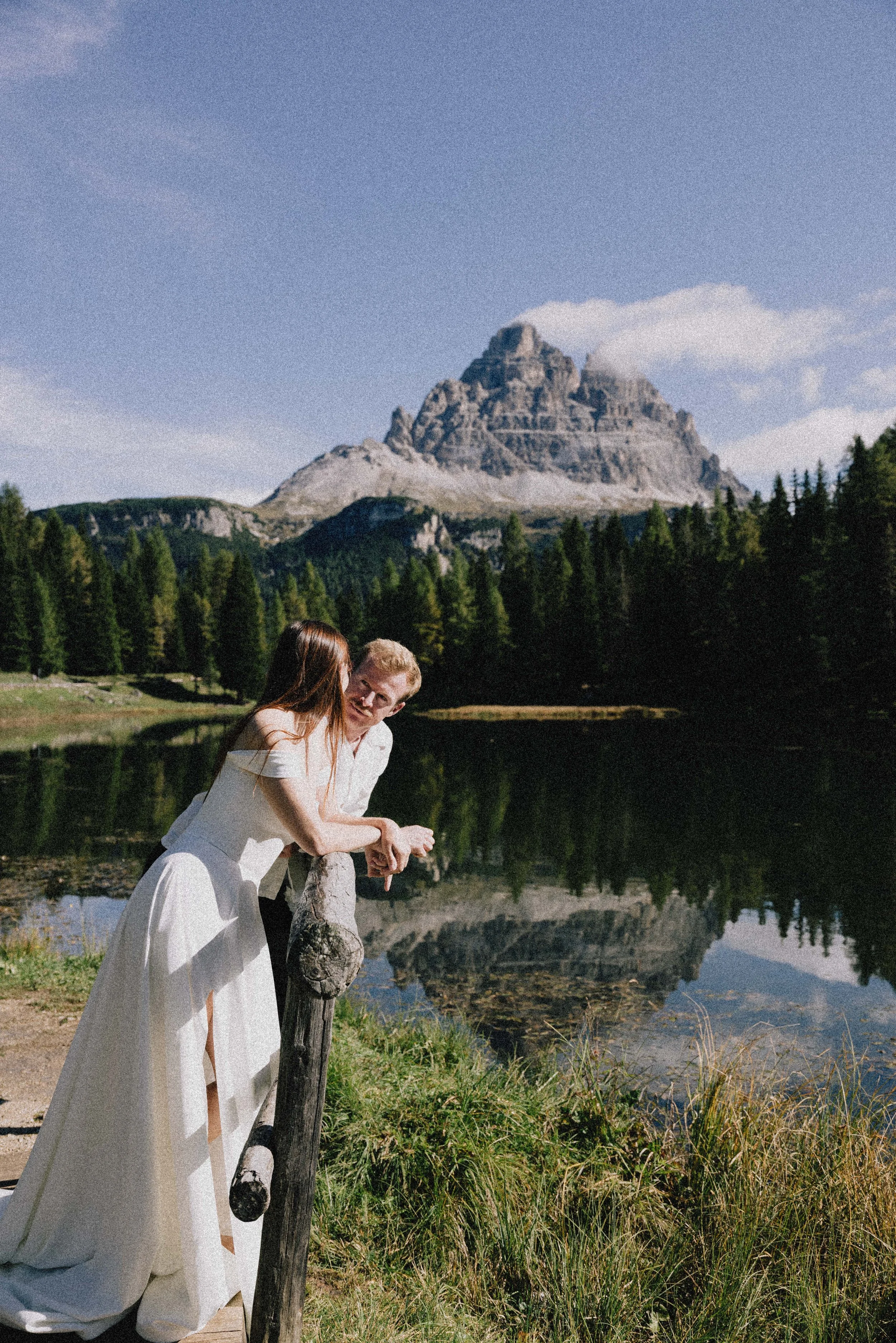 A couple in wedding attire leaning on a post by a lakeshore, with a large mountain and pine trees in the background.