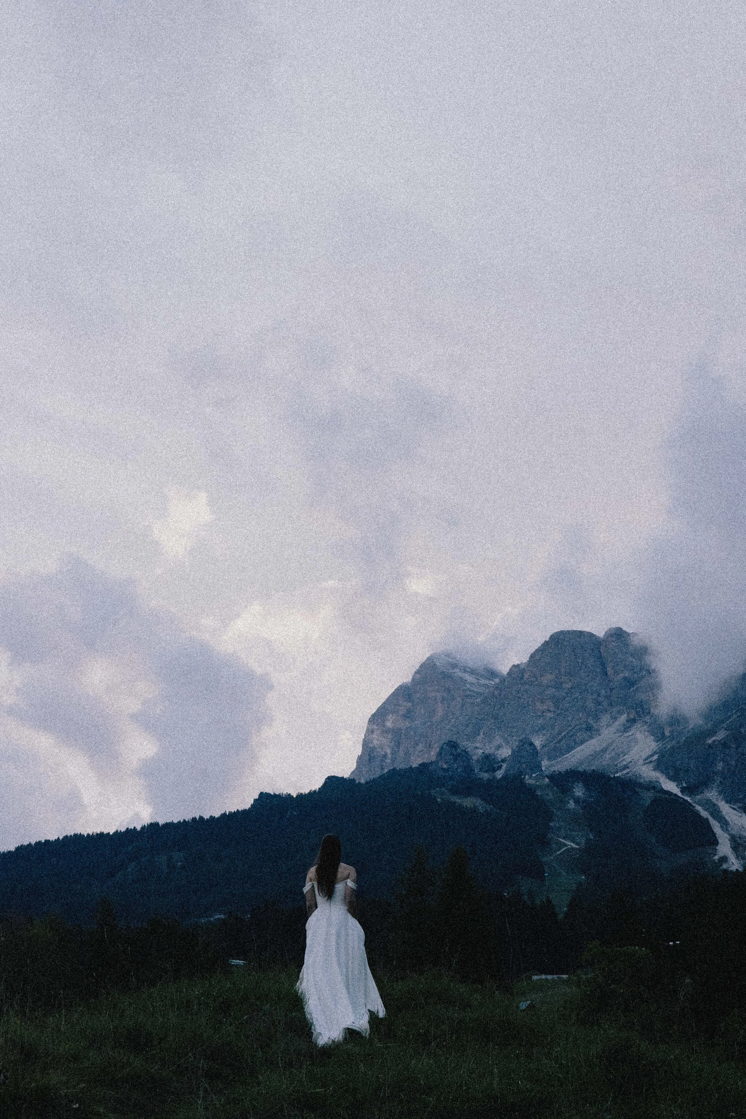 A woman in a white dress standing on green grass, looking at a mountain range with snow-capped peaks and cloudy sky in the background.
