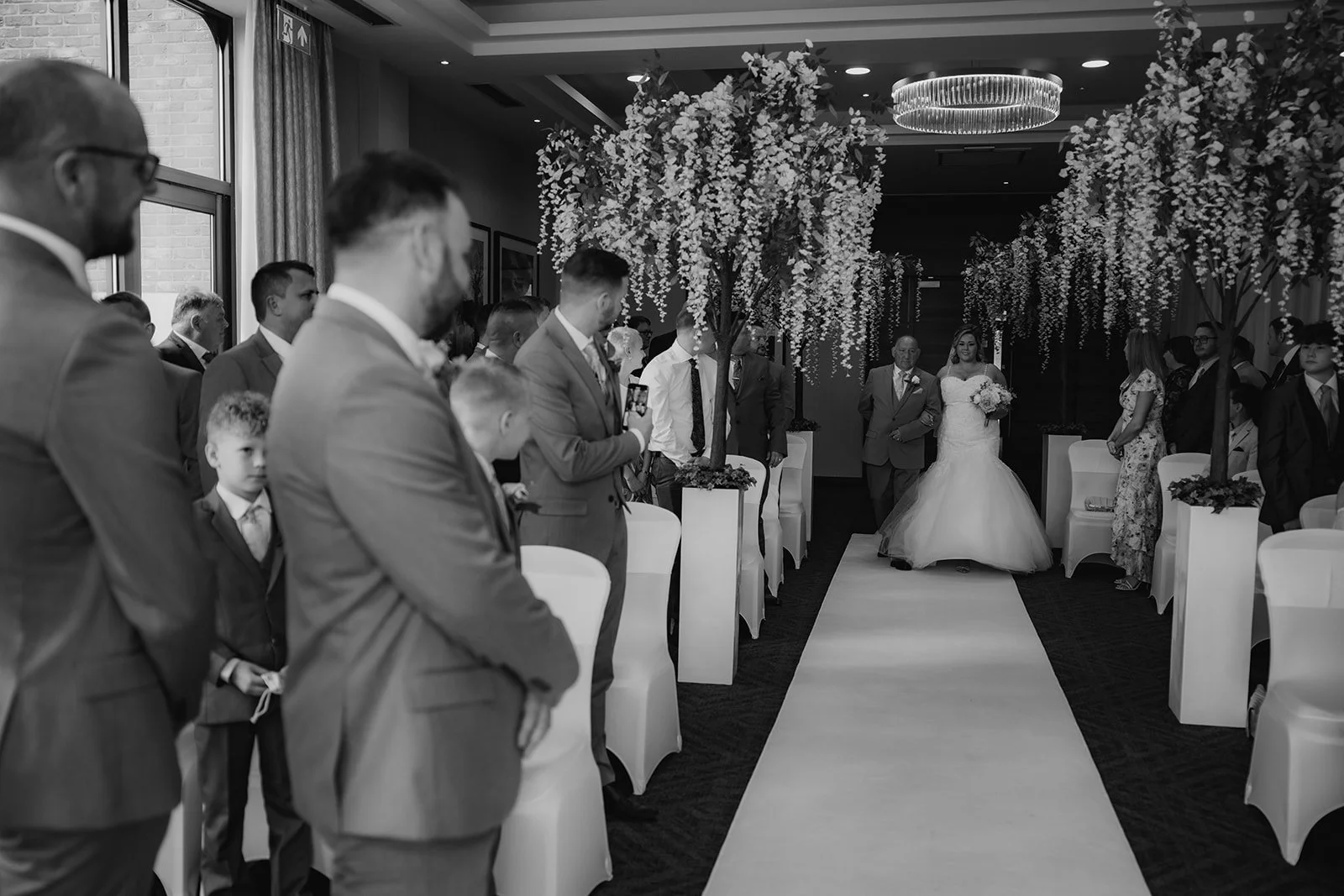 A wedding ceremony in an indoor venue with floral decorations hanging from the ceiling, guests standing on either side of an aisle, and a bride walking down the aisle. Merrydale Manor Wedding Venue by documentary wedding photographer