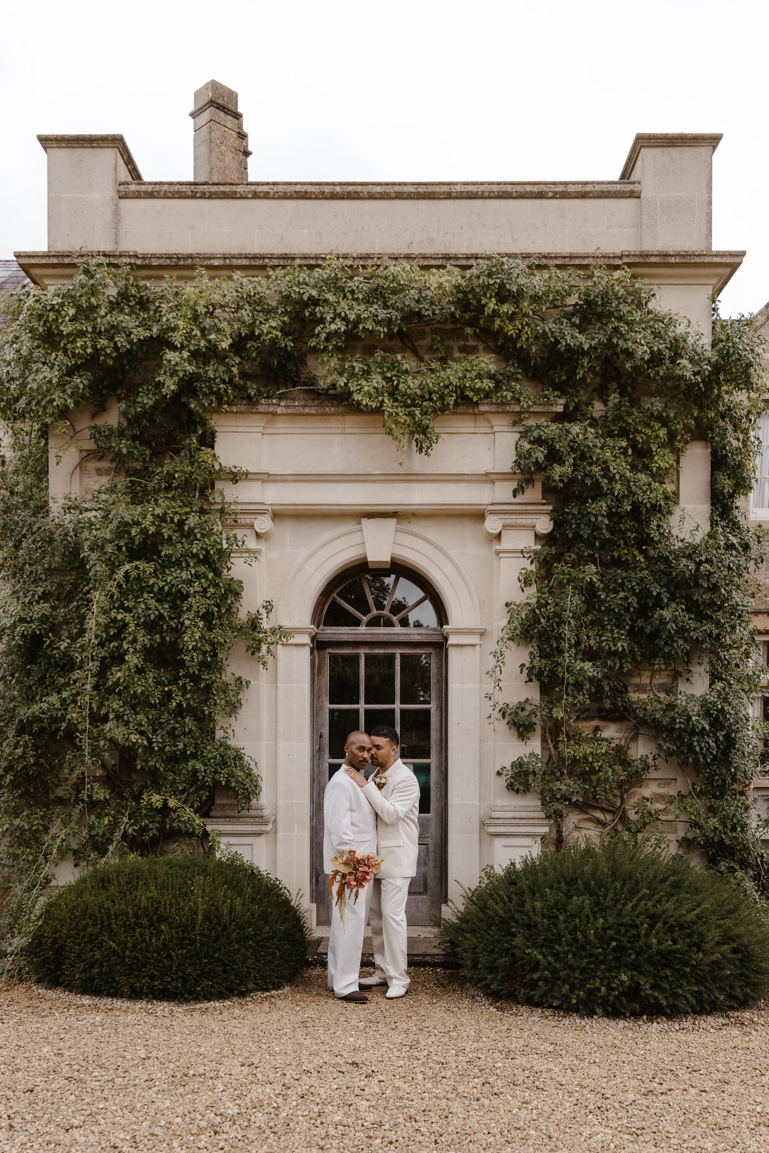 Two men in white suits standing close together in front of a historic building with green vines, holding a bouquet of flowers. LGBT wedding at euridge lodge 