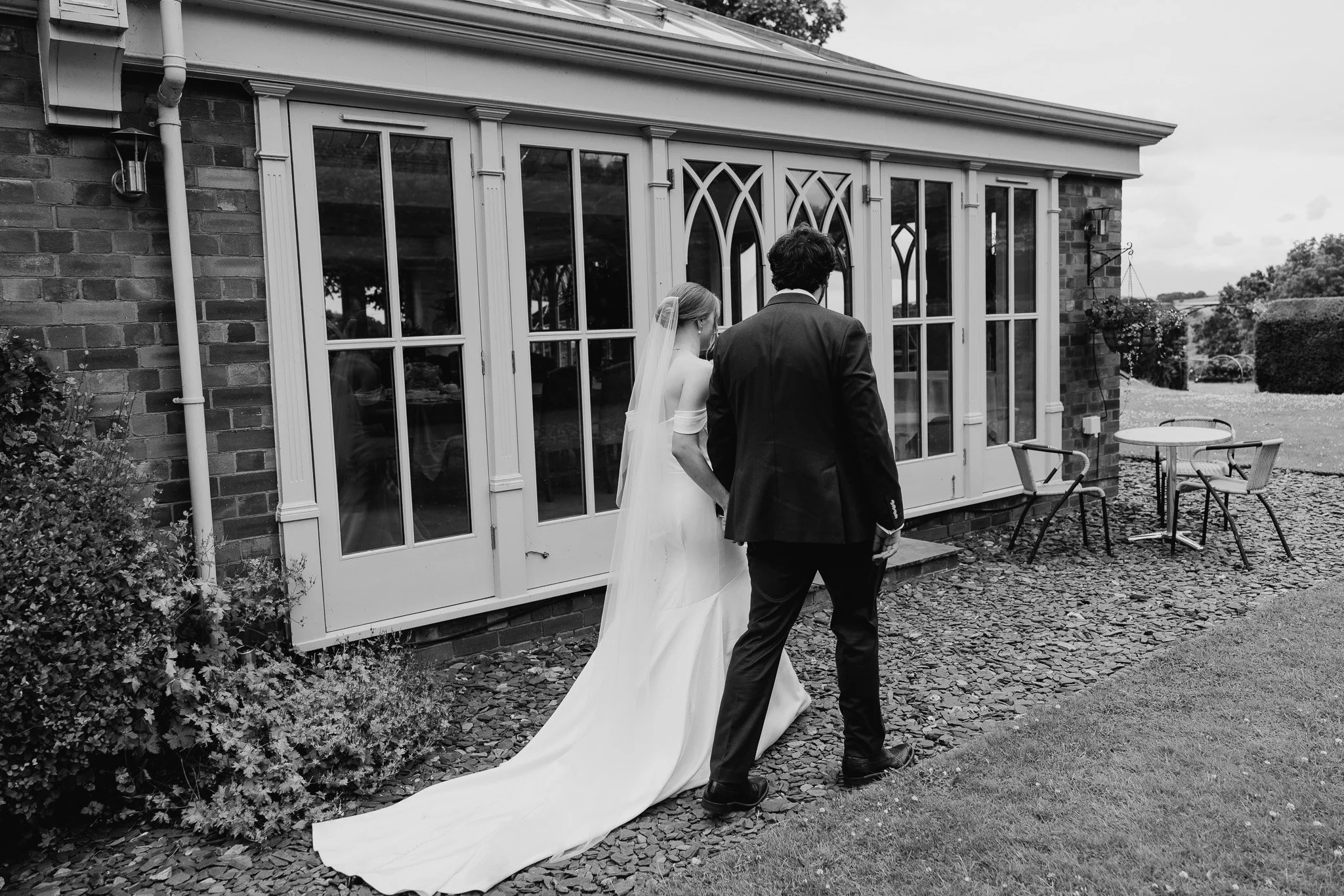 A bride in a wedding dress and veil walking with a groom in a suit on a stone-paved pathway outside a building with paneled glass doors.