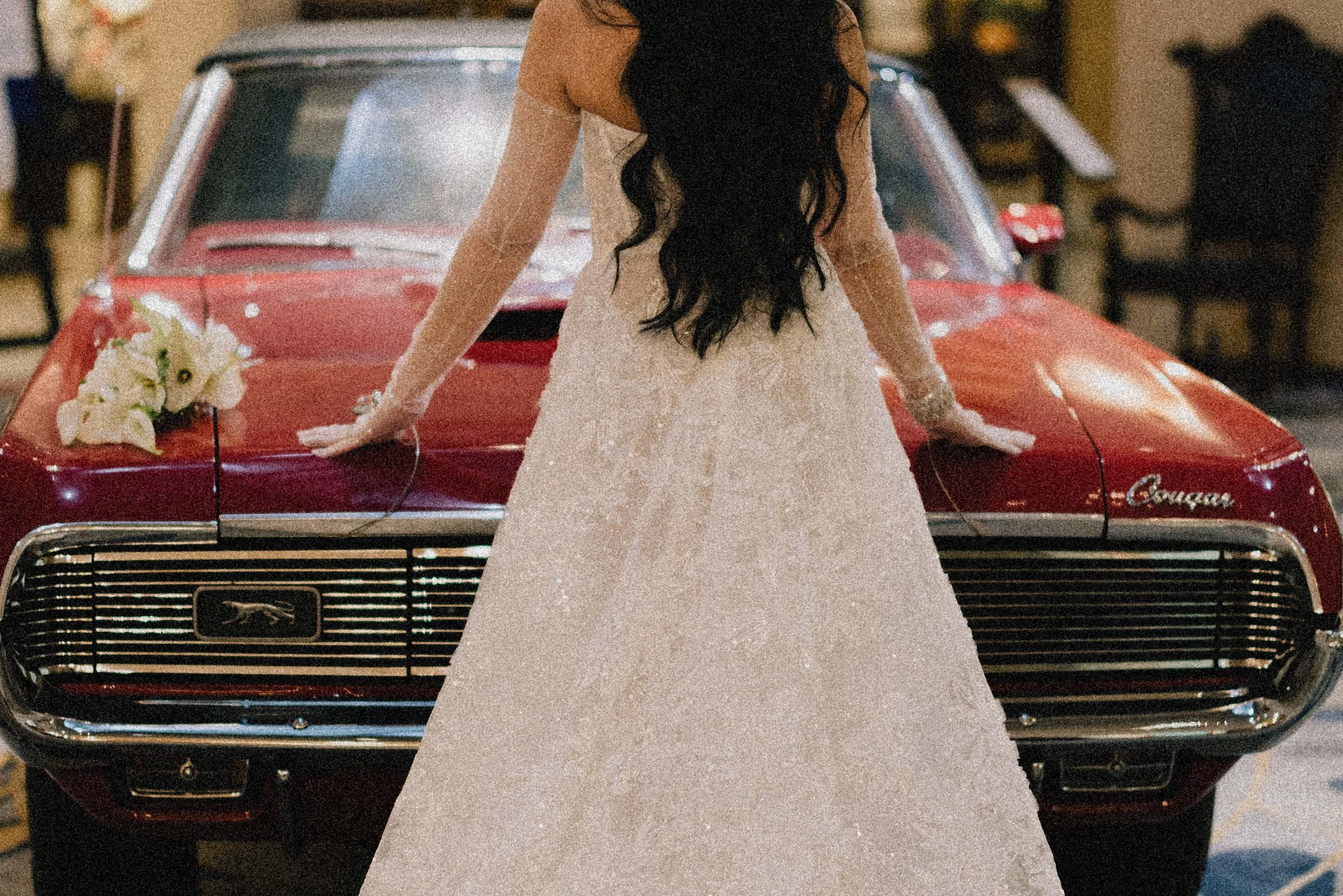 A bride in a white wedding dress with long dark hair, standing in front of a vintage red Cougar car, resting her hands on the car's hood, with a bouquet of white flowers placed on the car.