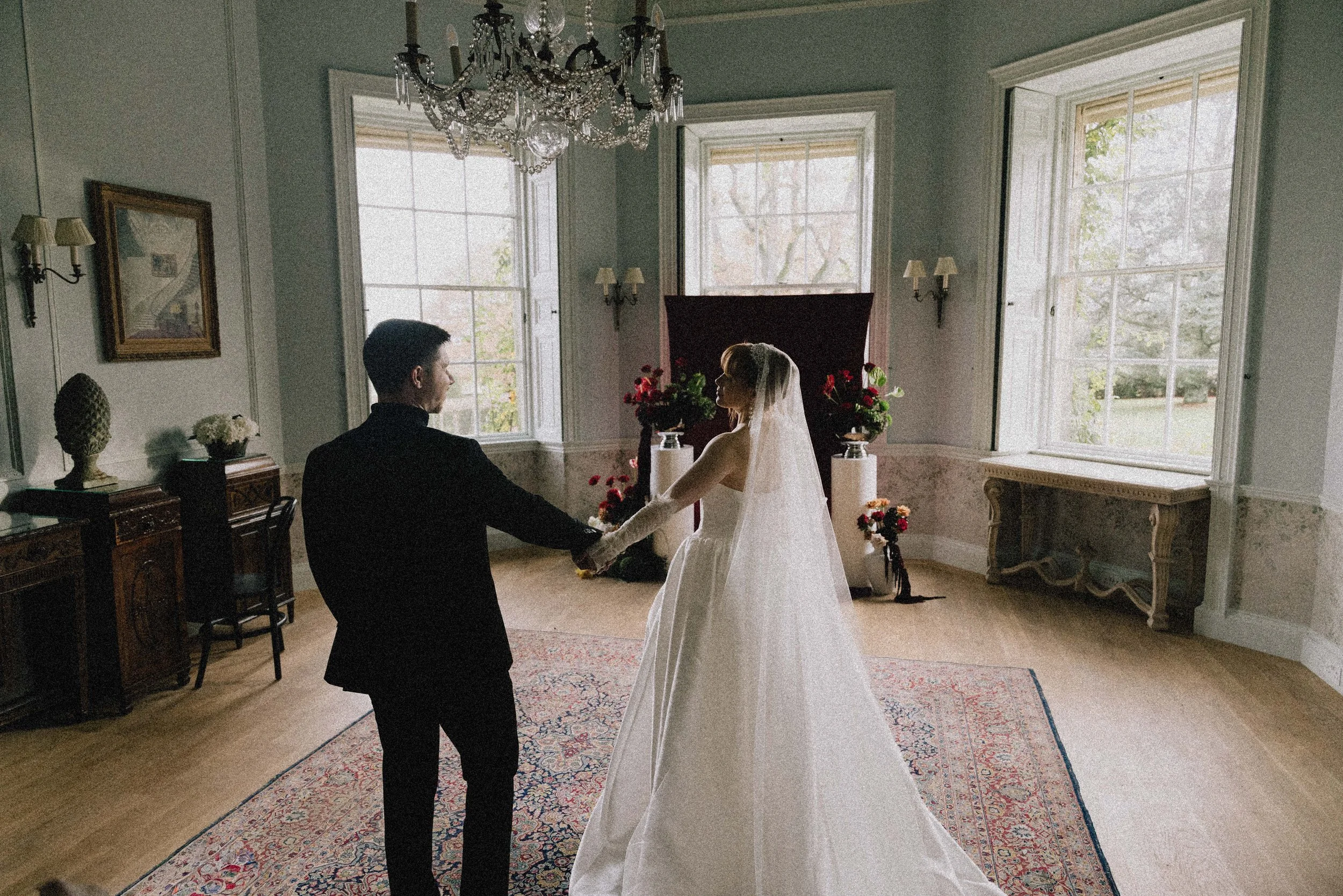 A bride and groom holding hands in a room with large windows, floral decor, and a chandelier, during their wedding ceremony.
