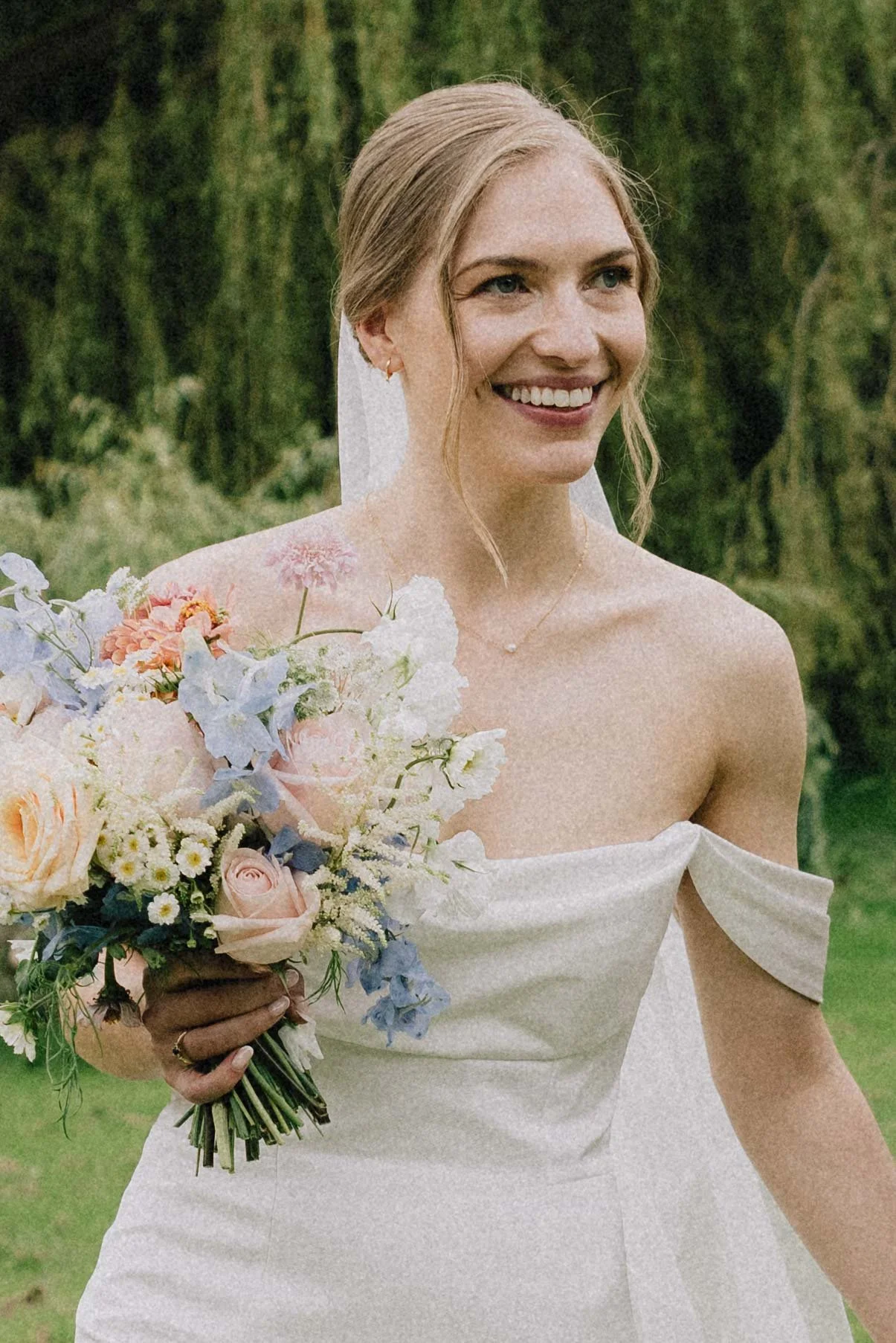 A smiling bride holding a bouquet of pastel-colored flowers outdoors with green trees in the background.