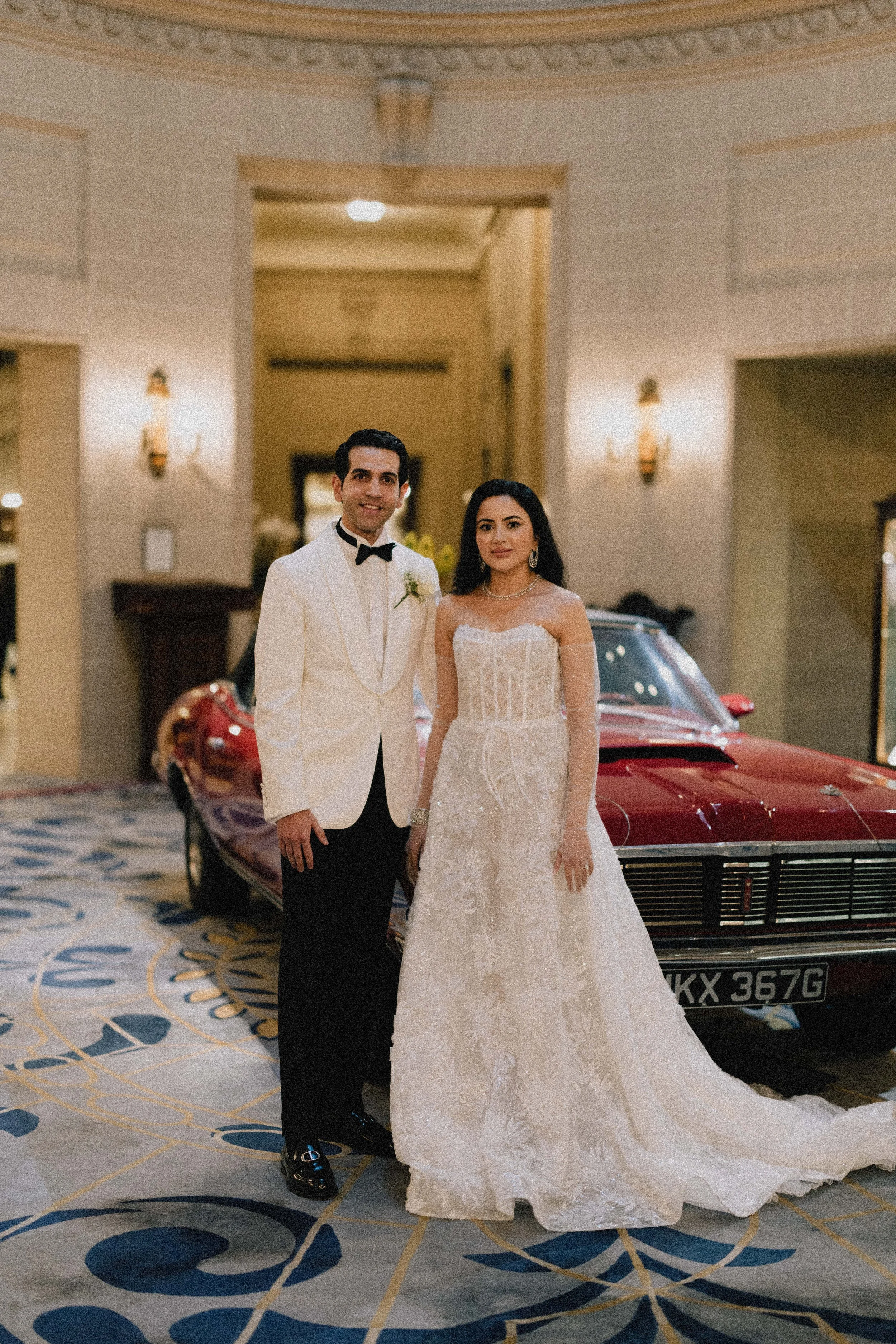 A bride and groom in wedding attire standing inside a grand hotel lobby with a vintage red sports car behind them. London wedding photography, RAC london wedding venue. Editorial wedding Photography 