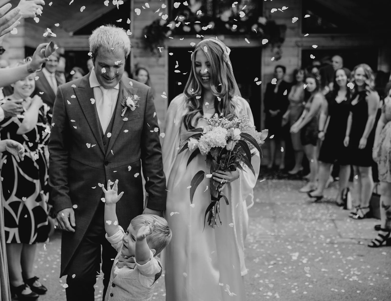 A wedding celebration with a bride, groom, and a young boy under falling confetti. The bride is holding a bouquet, and the groom is smiling. Guests are watching and smiling in the background. At wedding venue in Cheshire style lodge