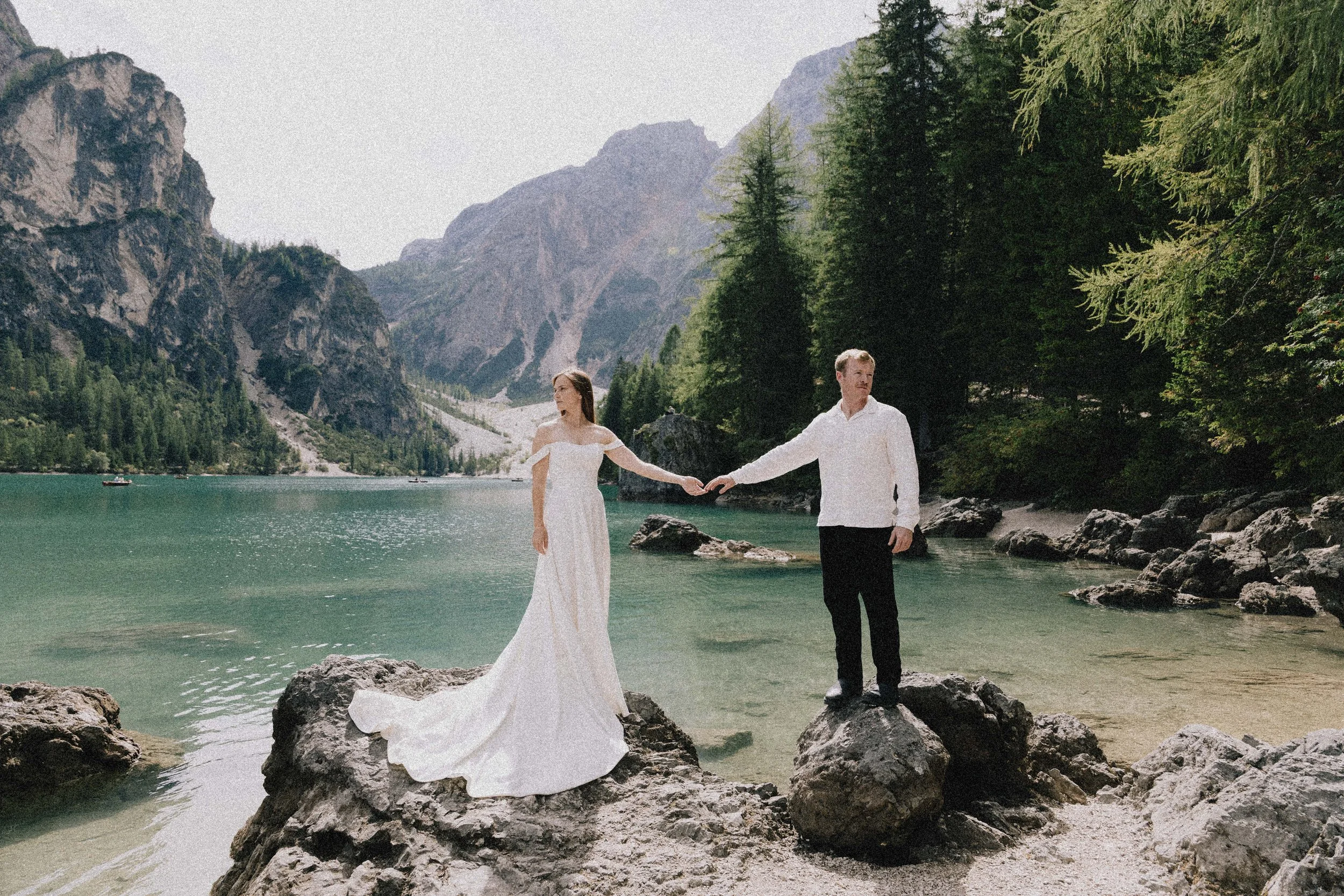 A couple in wedding attire standing hand in hand on rocks at the edge of a lake surrounded by mountains and forest.