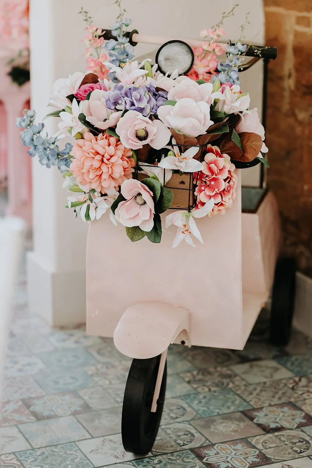 A bicycle with a front basket filled with colorful artificial flowers, including pink, purple, white, and blue blossoms, positioned indoors on patterned tile flooring. Merrydale Manor Wedding Venue