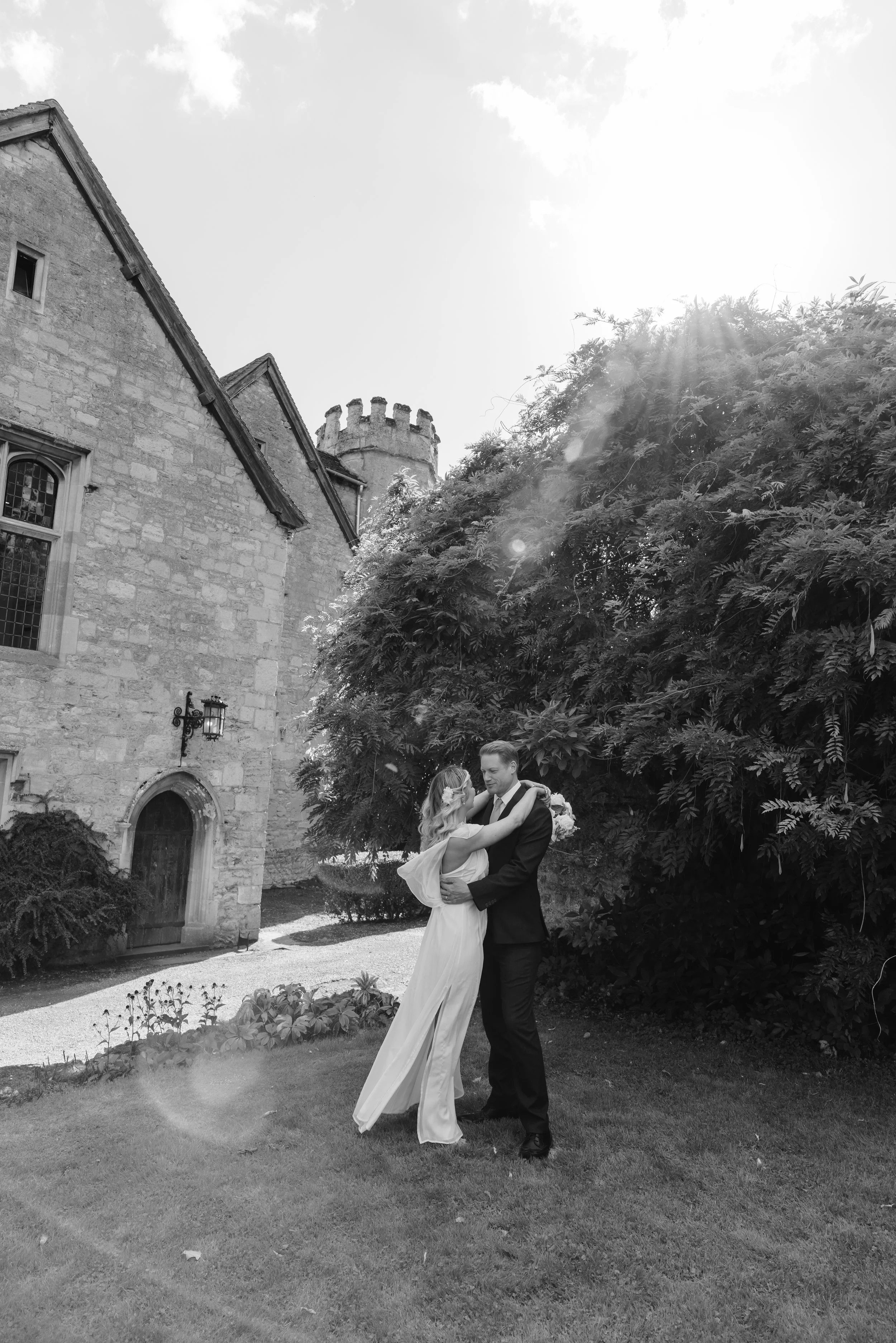 A newlywed couple in wedding attire embracing outdoors near a large bush and stone building with a tower, under bright sunlight. wedding in york, documentary wedding photographer
