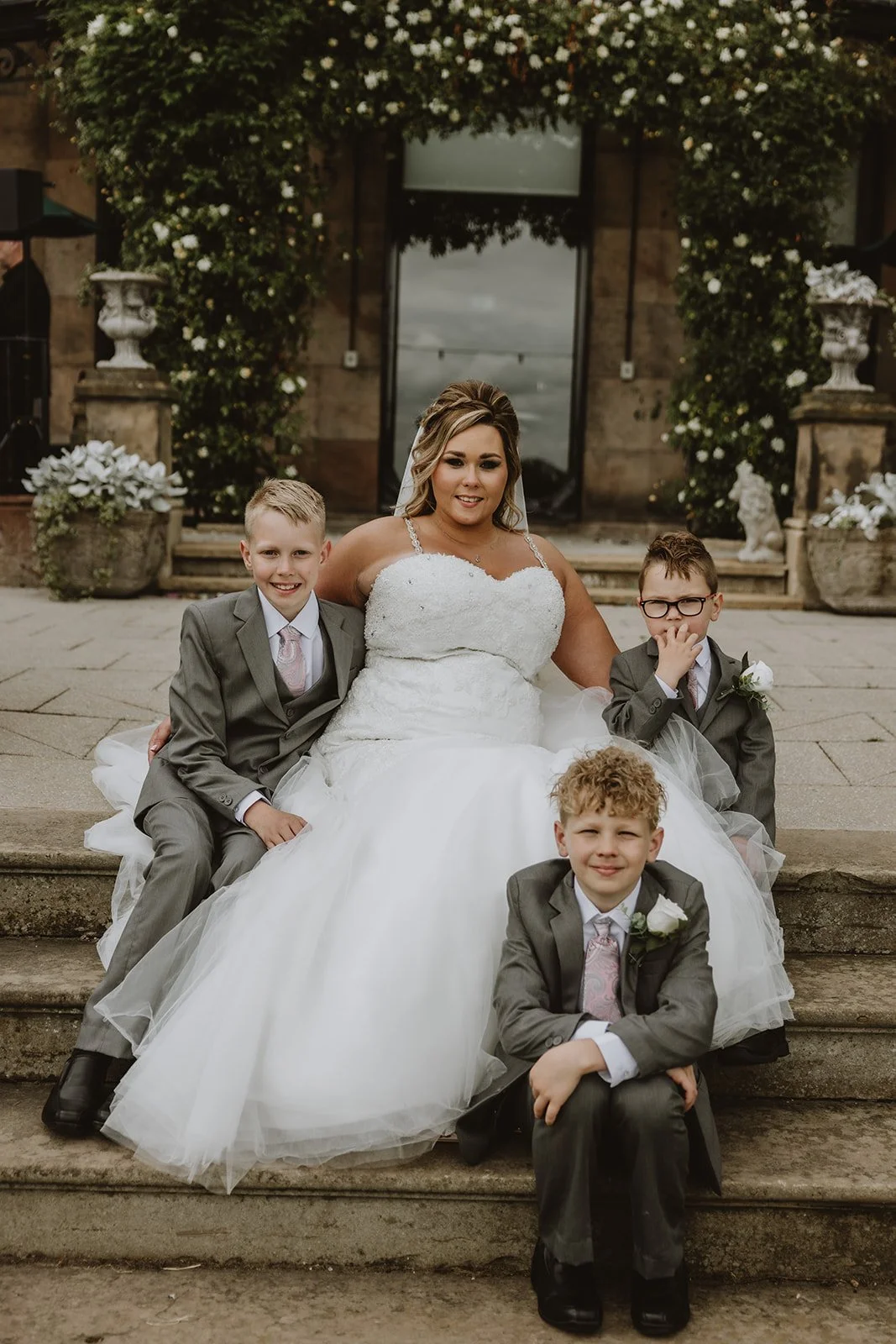 A bride in a white wedding dress sitting on stone steps with four young boys in gray suits and pink ties, outside a building adorned with white flowers and greenery. wedding at Merrydale Manor Wedding Venue in the uk, documentary wedding photographer