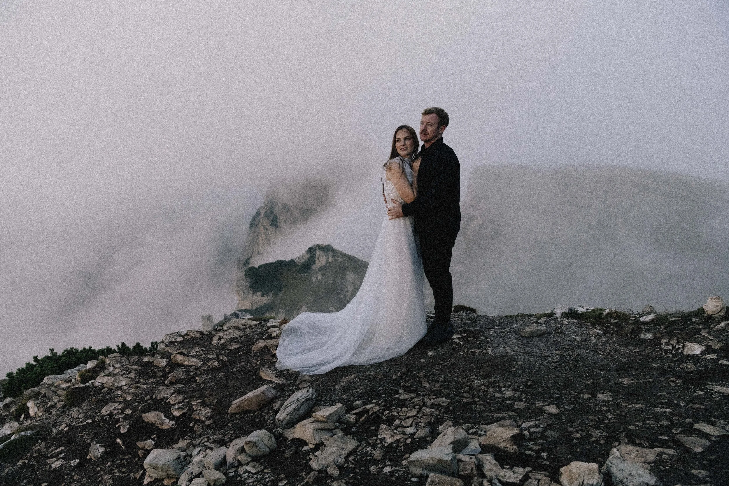 A couple dressed in wedding attire, standing on rocky terrain with clouds and mountains in the background.