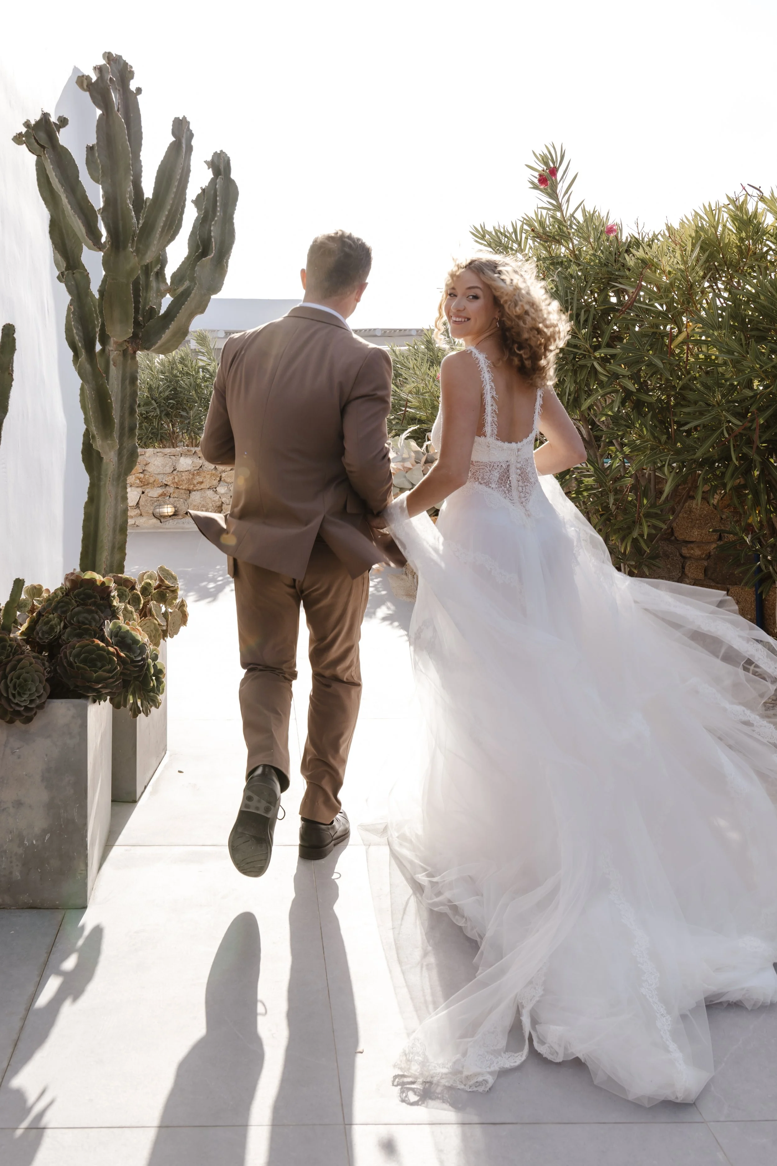 A bride and groom walking hand in hand outdoors, with the bride smiling at the camera, surrounded by desert plants including a large cactus and succulents, during sunset. Uk destination wedding photographer in Greece