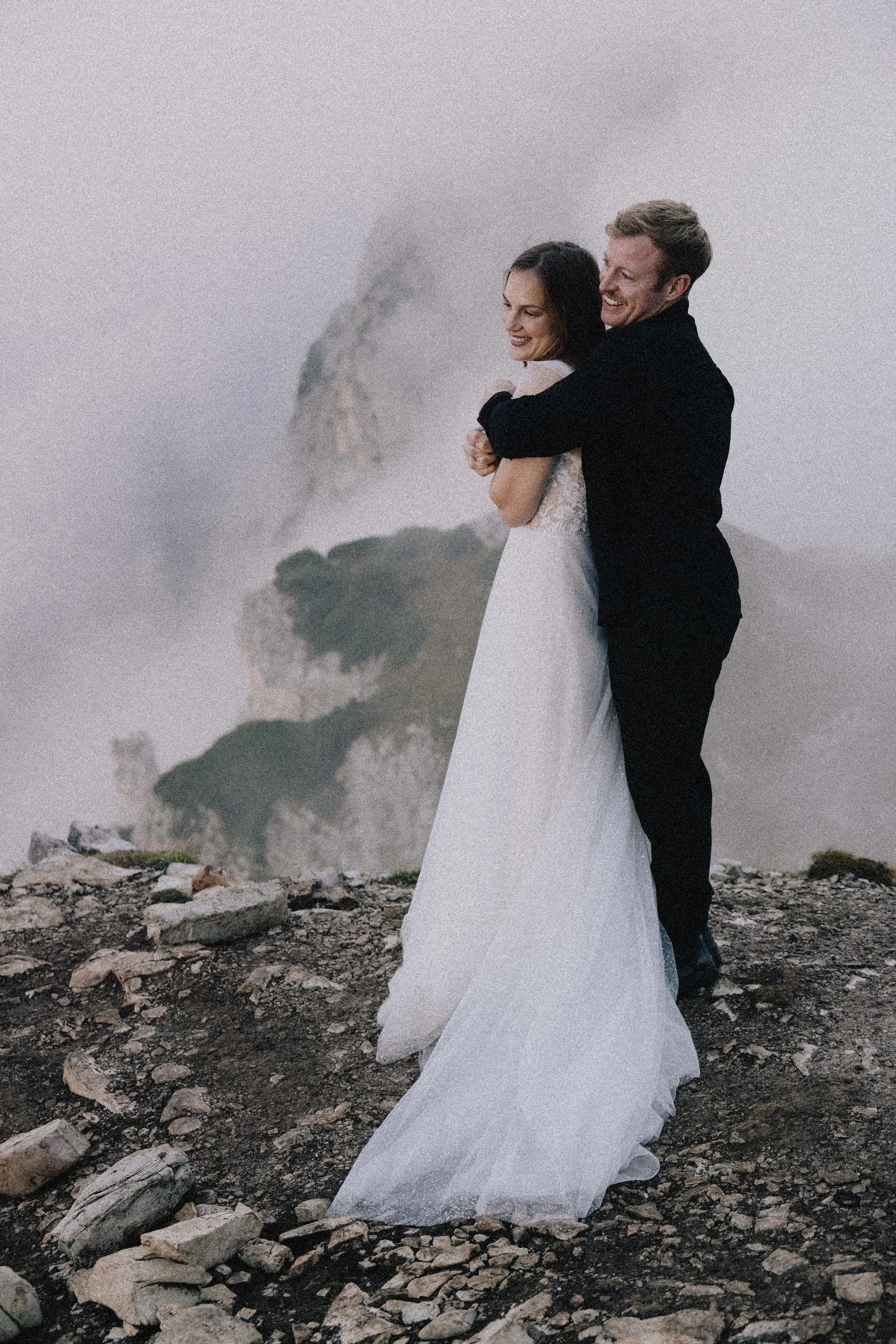 A newlywed couple standing on rocky terrain embracing each other with a misty mountain in the background.