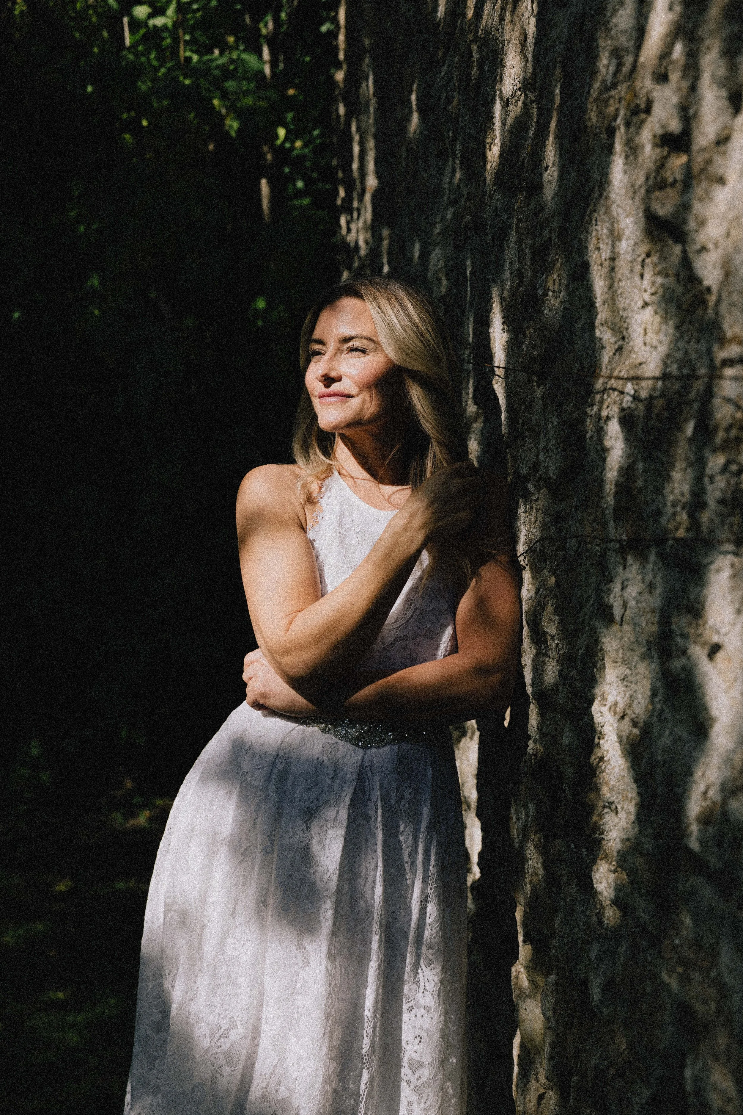 A woman in a white lace dress standing next to a large stone wall, with sunlight illuminating her face and upper body.