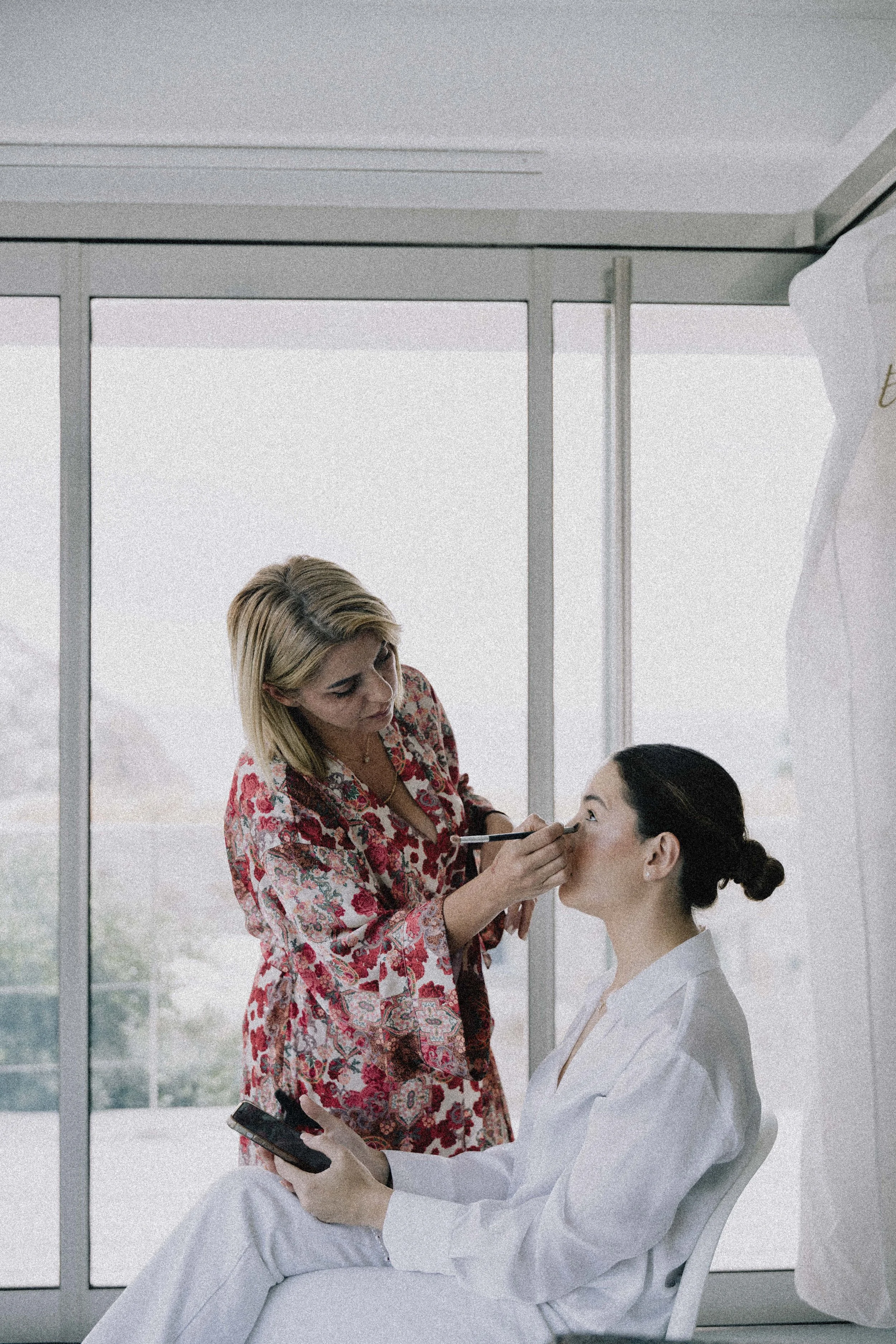A woman in a colorful blouse applies makeup to another woman in a white outfit, sitting in front of a large window.