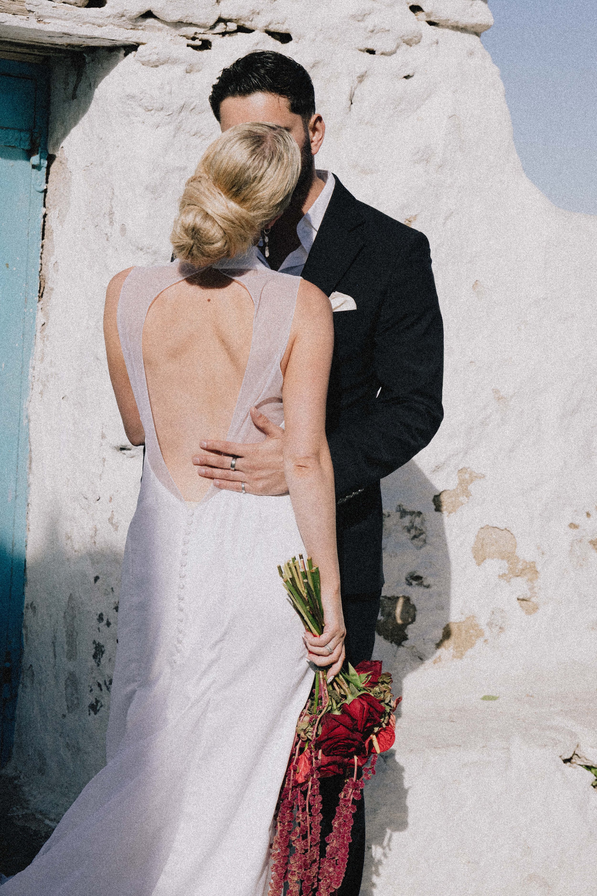 A bride and groom sharing a kiss outdoors, with the bride holding a bouquet of red flowers, against a white, textured stone wall.