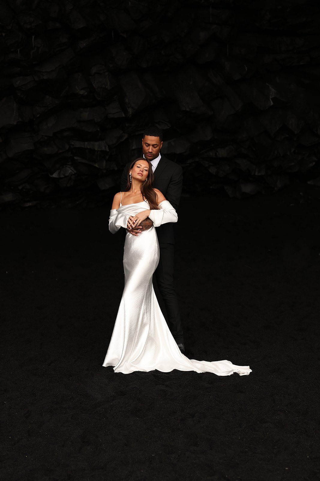 A couple dressed in wedding attire standing in front of a dark, rocky background. The woman in a white gown and the man in a black suit embrace each other. documentary wedding photographer for iceland elopment
