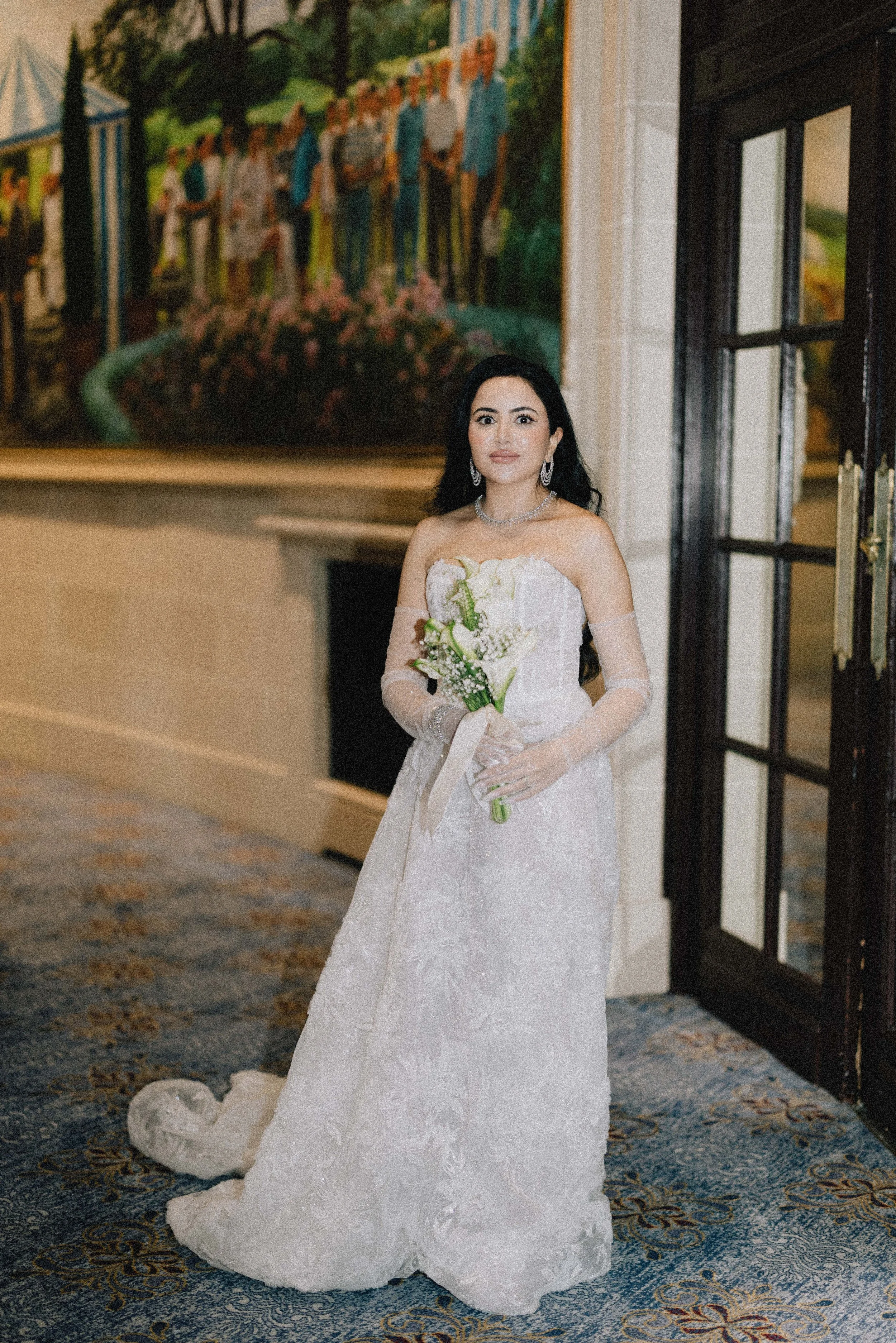 A bride in a strapless white wedding gown holding a bouquet of white flowers, standing indoors with a large colorful painting in the background. london wedding venue 