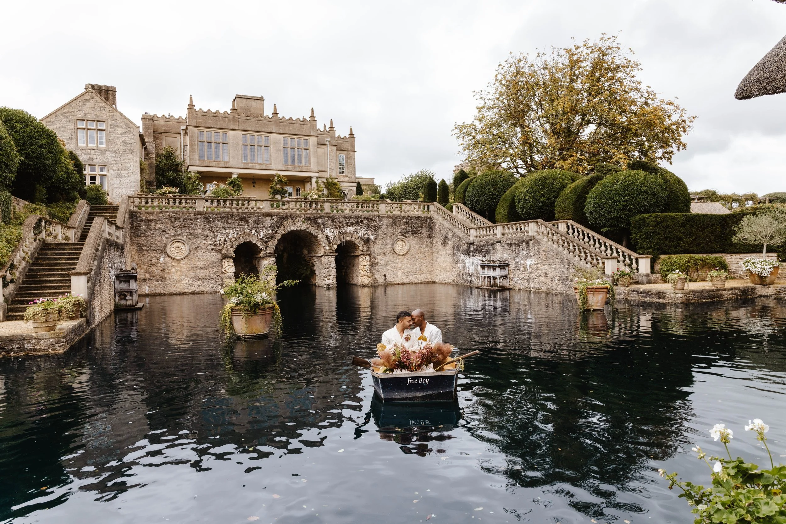 Two people in a boat decorated with flowers on a waterway with historic stone buildings and manicured trees in the background. weddings at euridge manor 