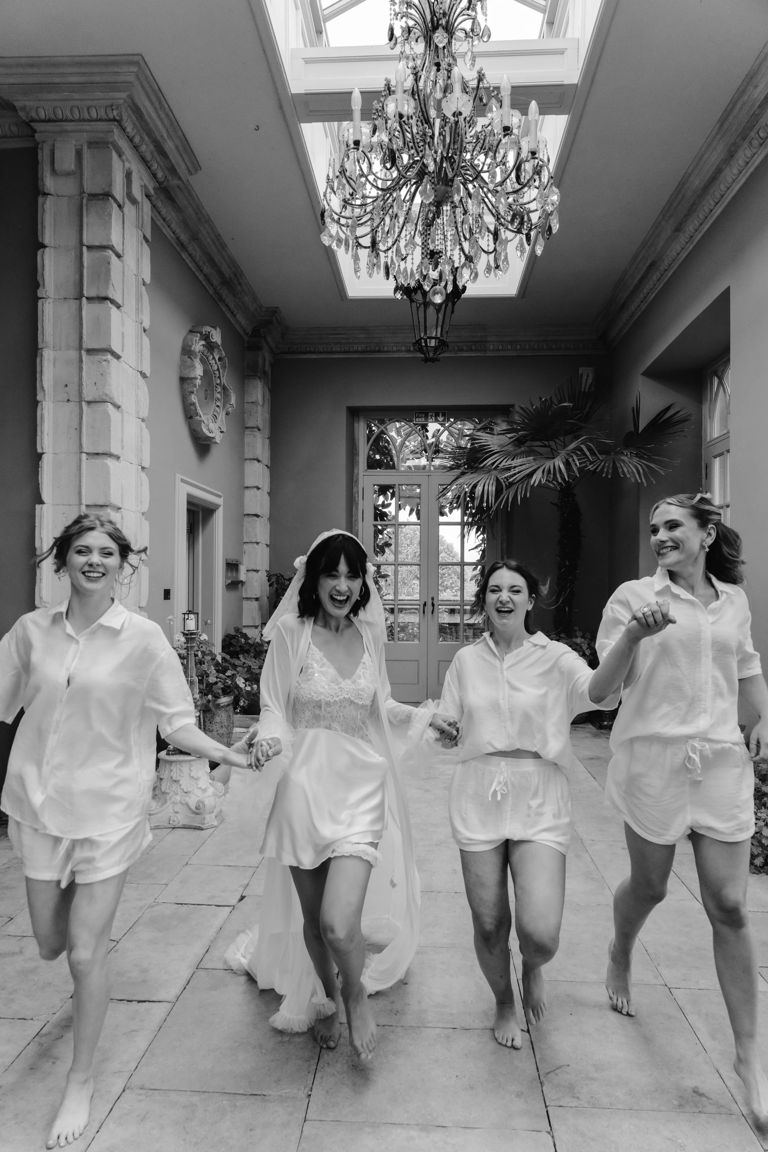 A bride and three women in sleepwear running and laughing indoors beneath a chandelier and skylight. wedding at euridge manor 